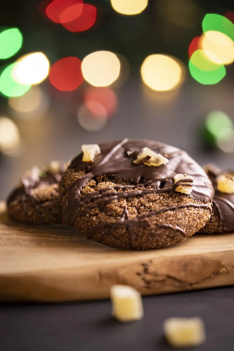 Close-up of chocolate ginger cookies with ginger chunks on a wooden board, with colorful blurred holiday lights in the background.
