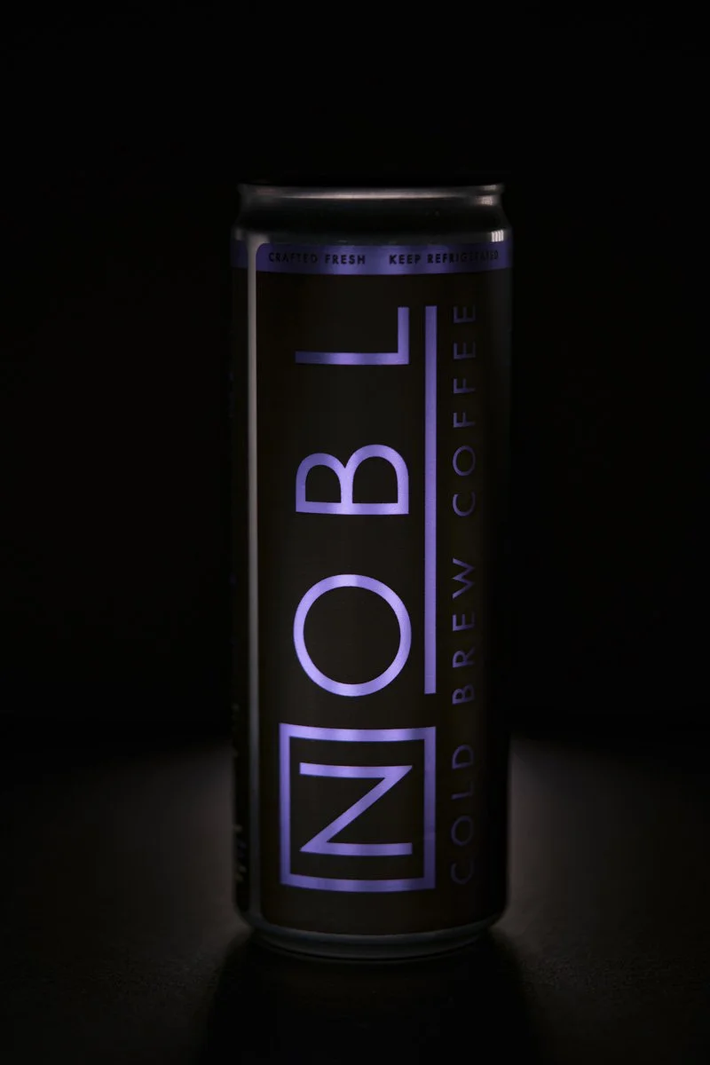 A tall aluminum can of NOBL cold brew coffee, illuminated with purple light, against a dark background.