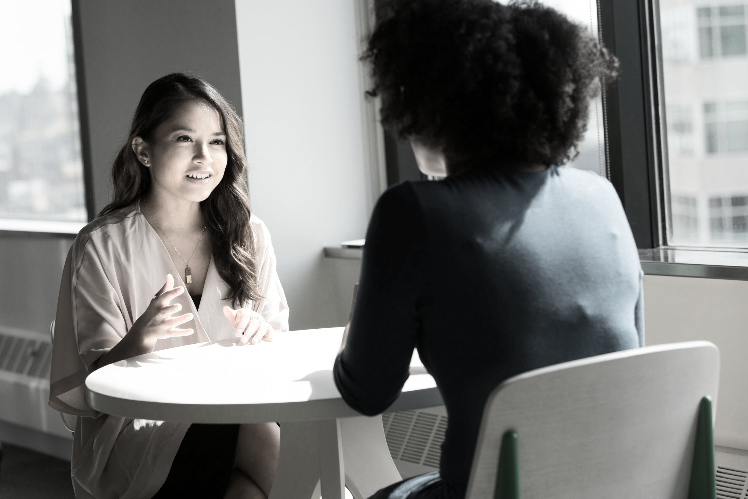 Two women having a conversation at a table by a large window in a modern office setting, with natural light.