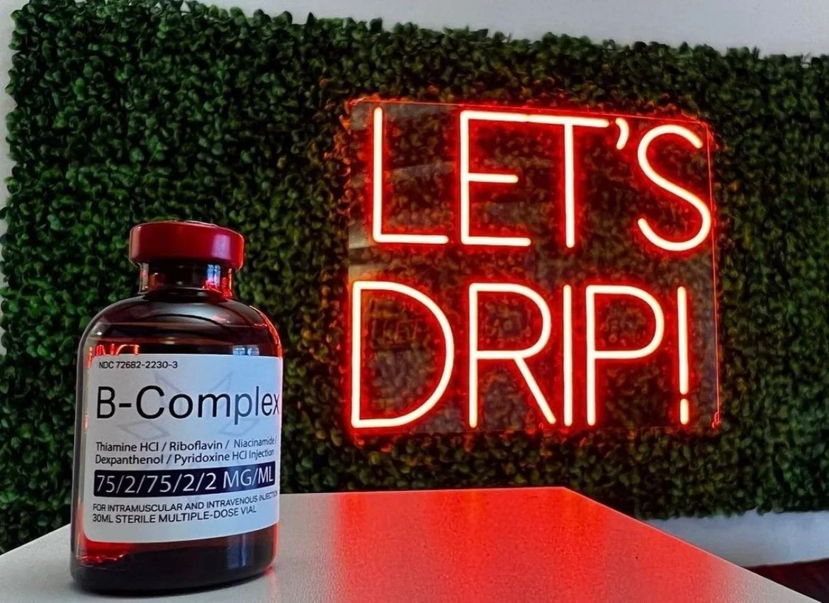 A bottle of B-Complex vitamins placed on a table in front of a wall with green foliage and a red neon sign that reads "LET'S DRIP!"