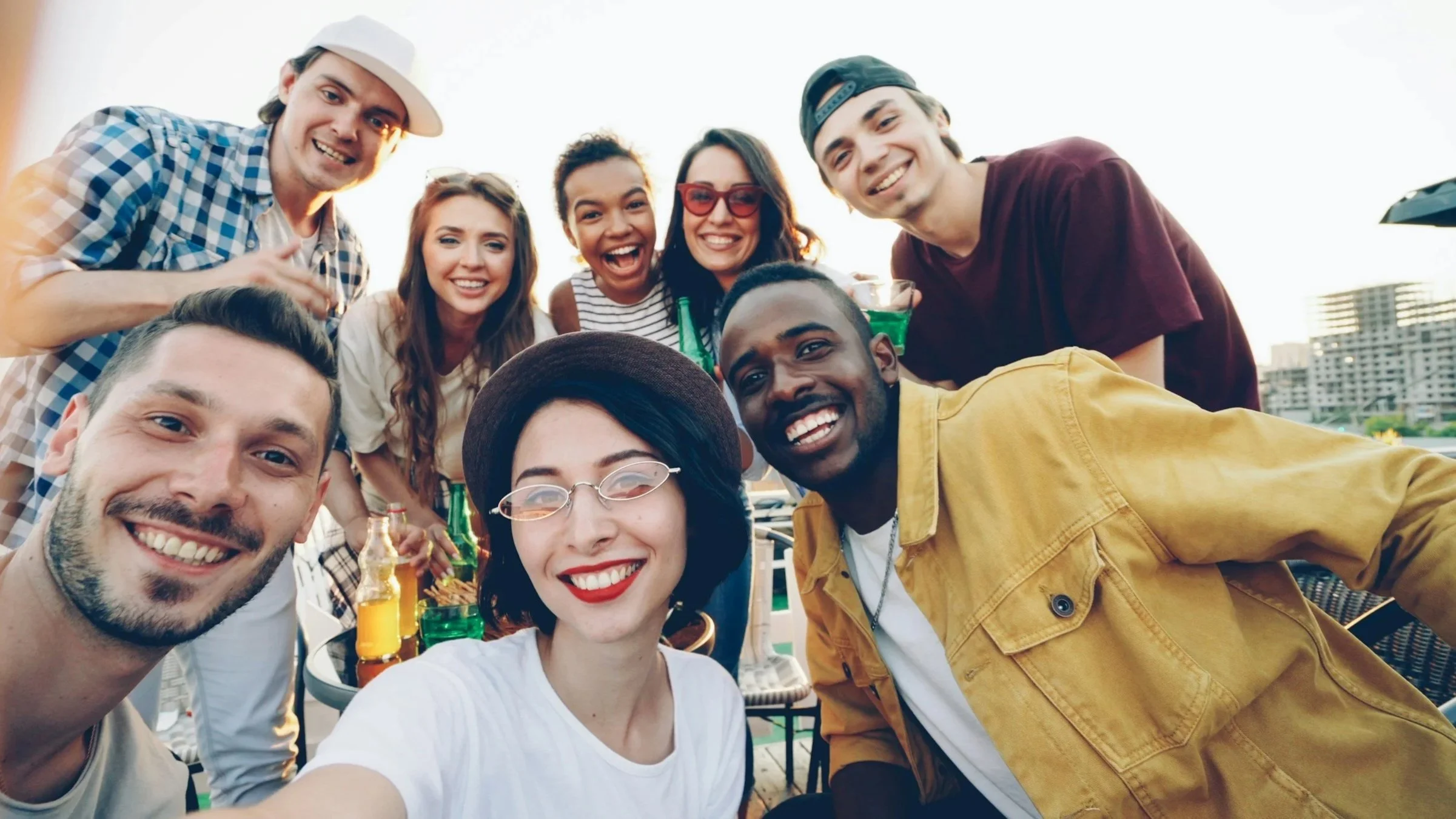 Group of friends smiling and taking a selfie at a rooftop party with city buildings in the background, enjoying drinks and good weather.