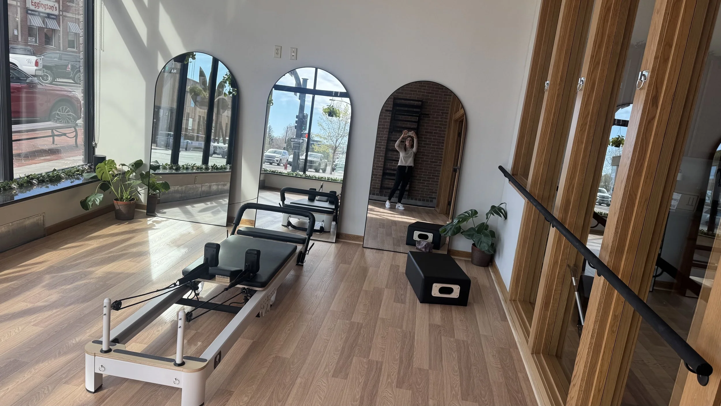 A fitness or physical therapy room with light wood flooring, a treatment or therapy table, a mirror, plants, and large windows letting in natural light. A person is taking a photo in the mirror.
