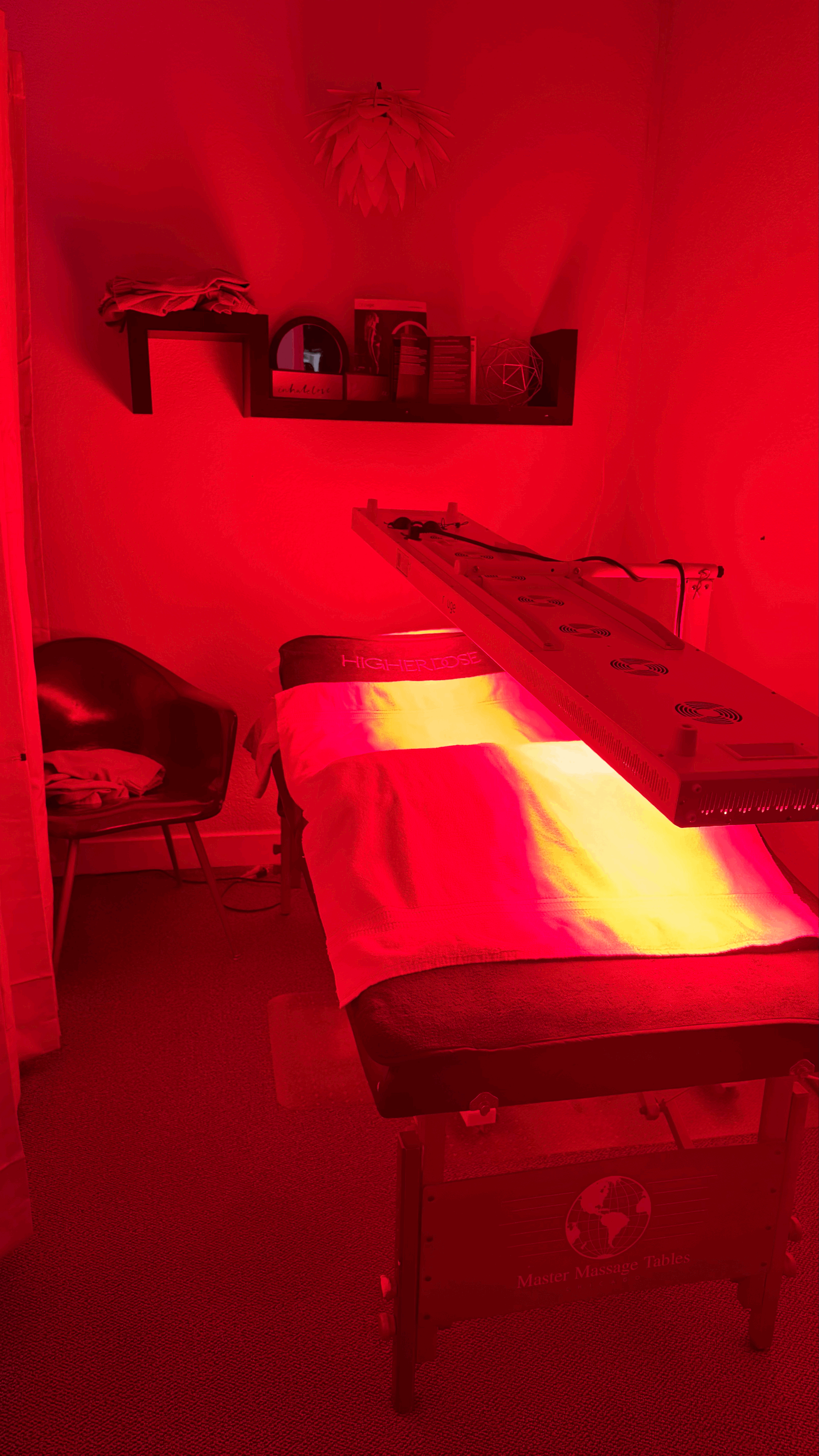 A massage therapy room with a massage table under red light, a shelf with decor and books on the wall, a black chair with clothes, and a red light therapy device over the massage table.