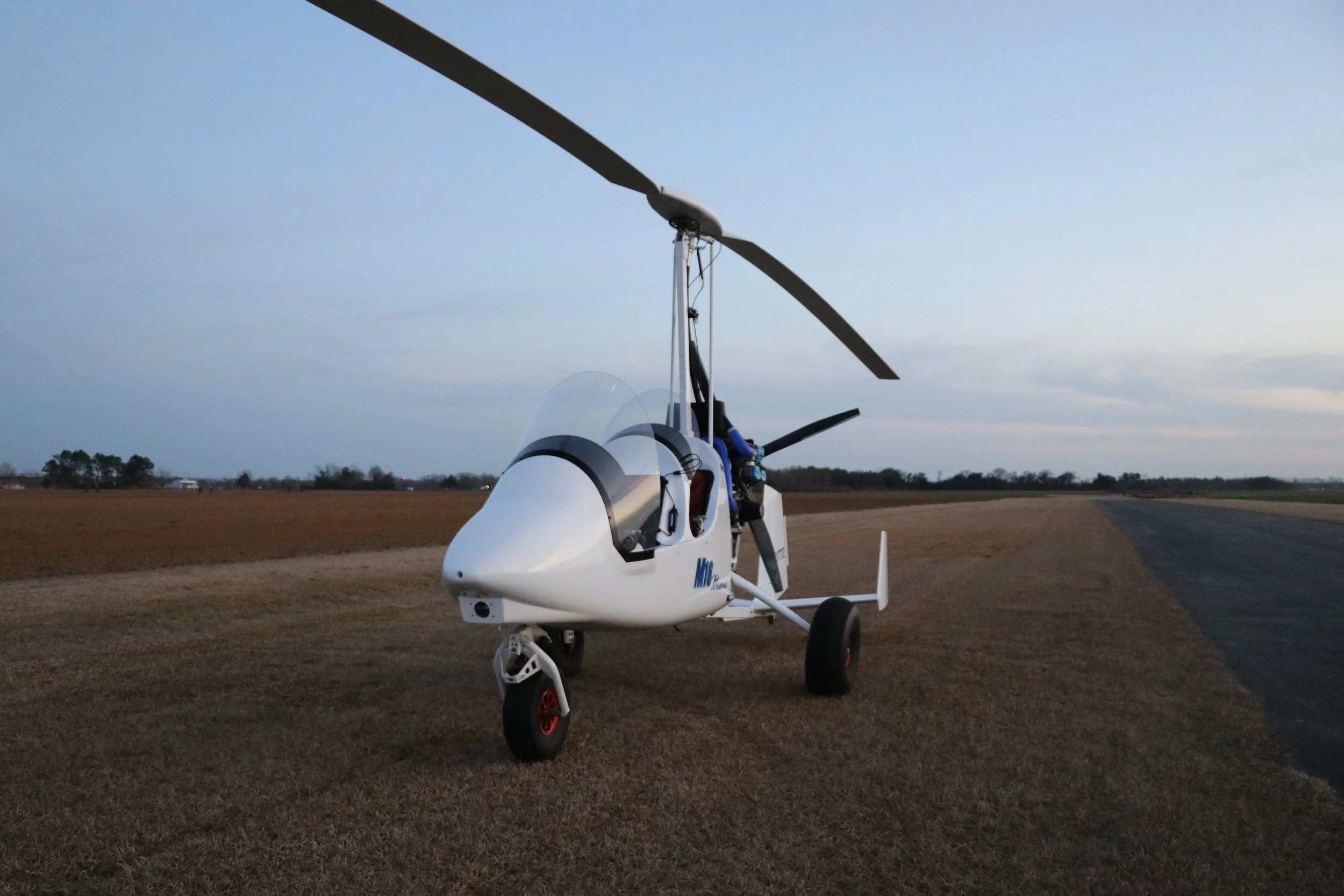 A white M16 gyroplane parked on a grassy field near a paved runway, with a cloudy sky in the background.