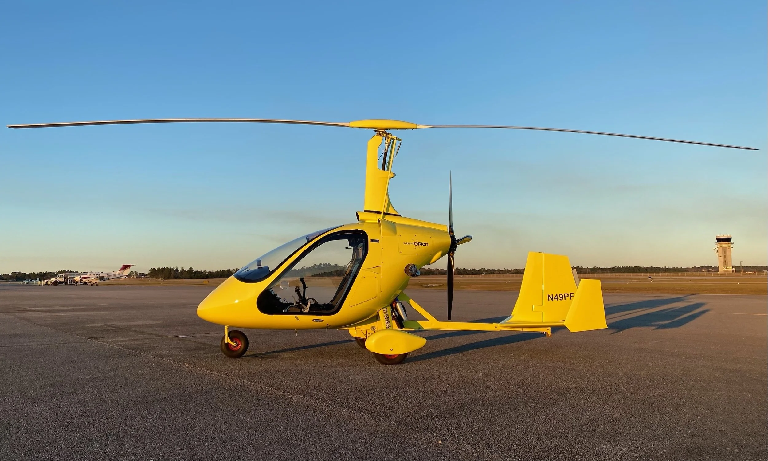 Yellow M24 parked on an airport tarmac with a control tower and small aircraft in the background at dusk.