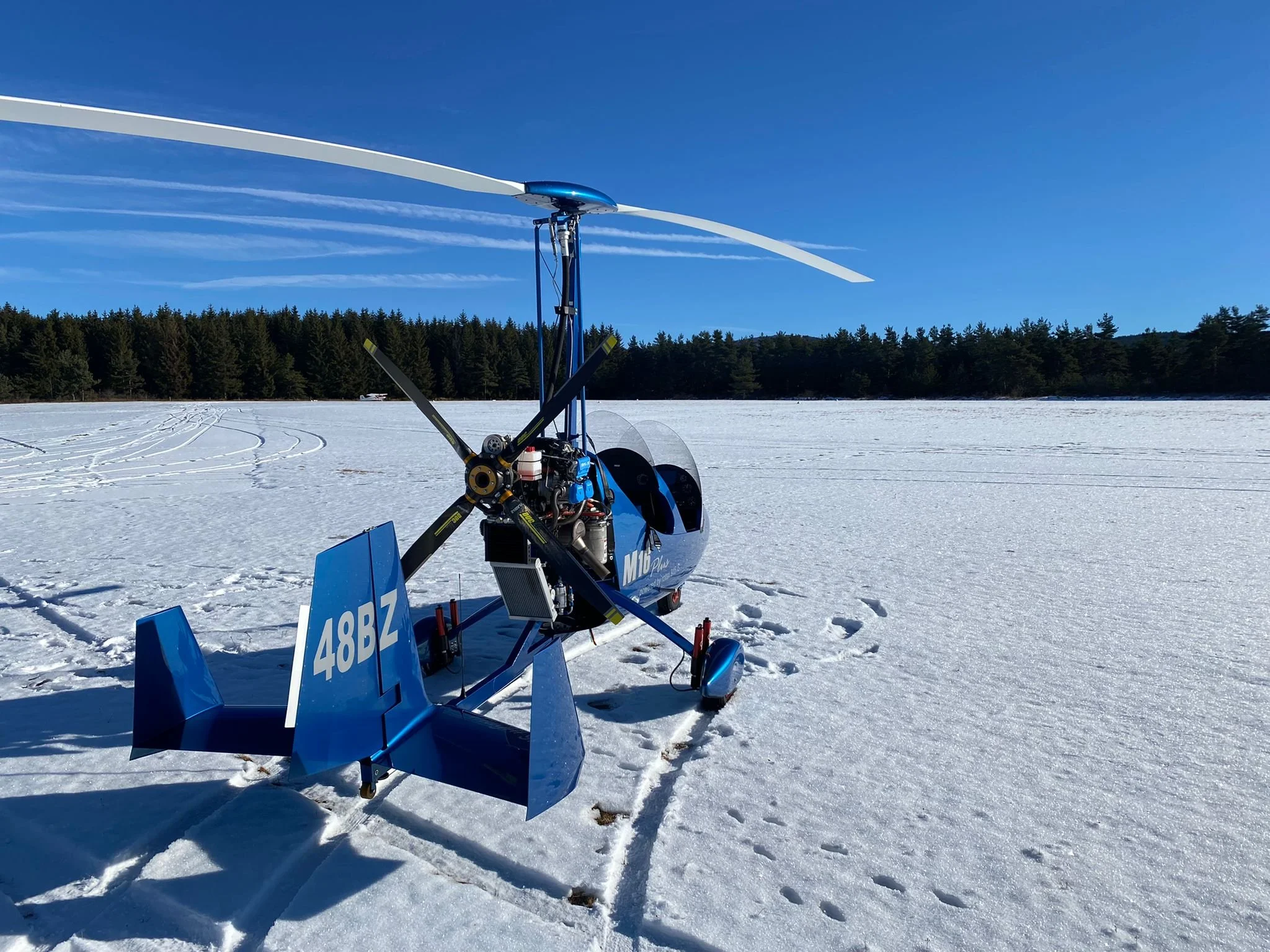 A blue gyroplane parked on snow-covered ground in an open, snowy field with trees in the background under a clear blue sky.