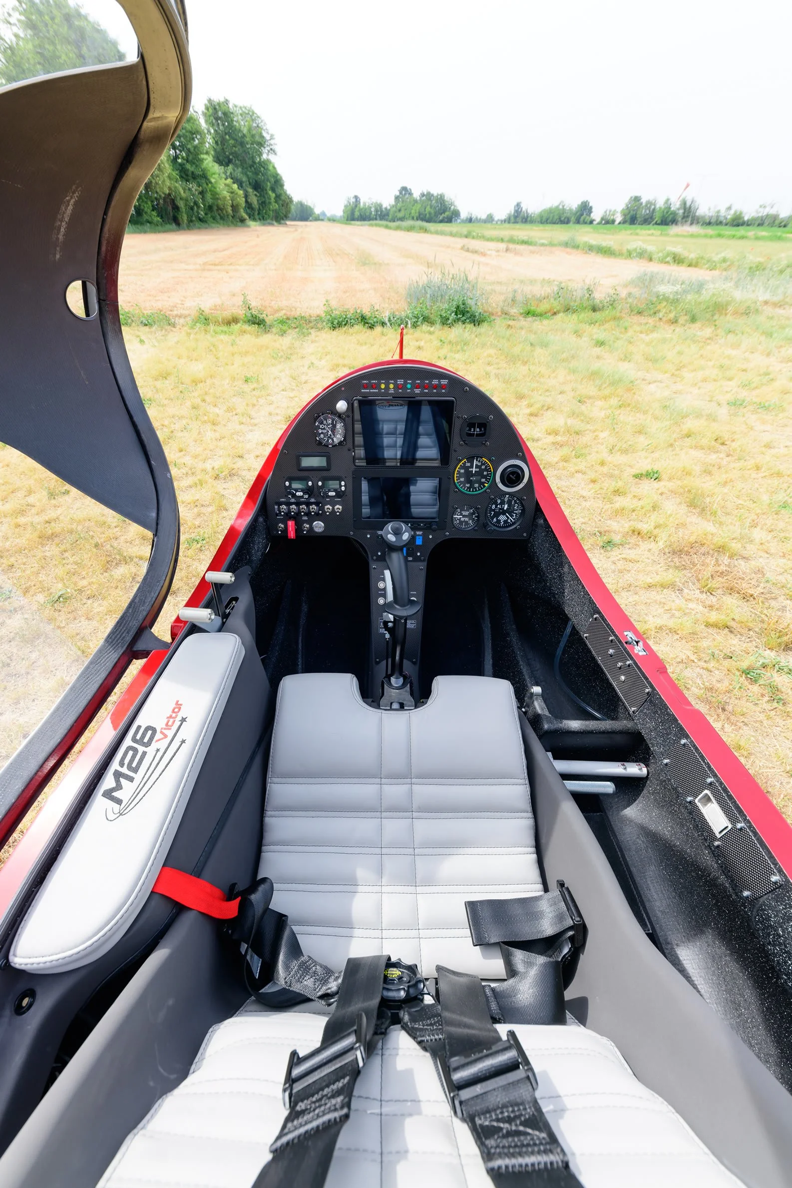 Inside view of a red M26 gyroplane, with a view of the grassy field ahead and various instruments and controls on the panel.
