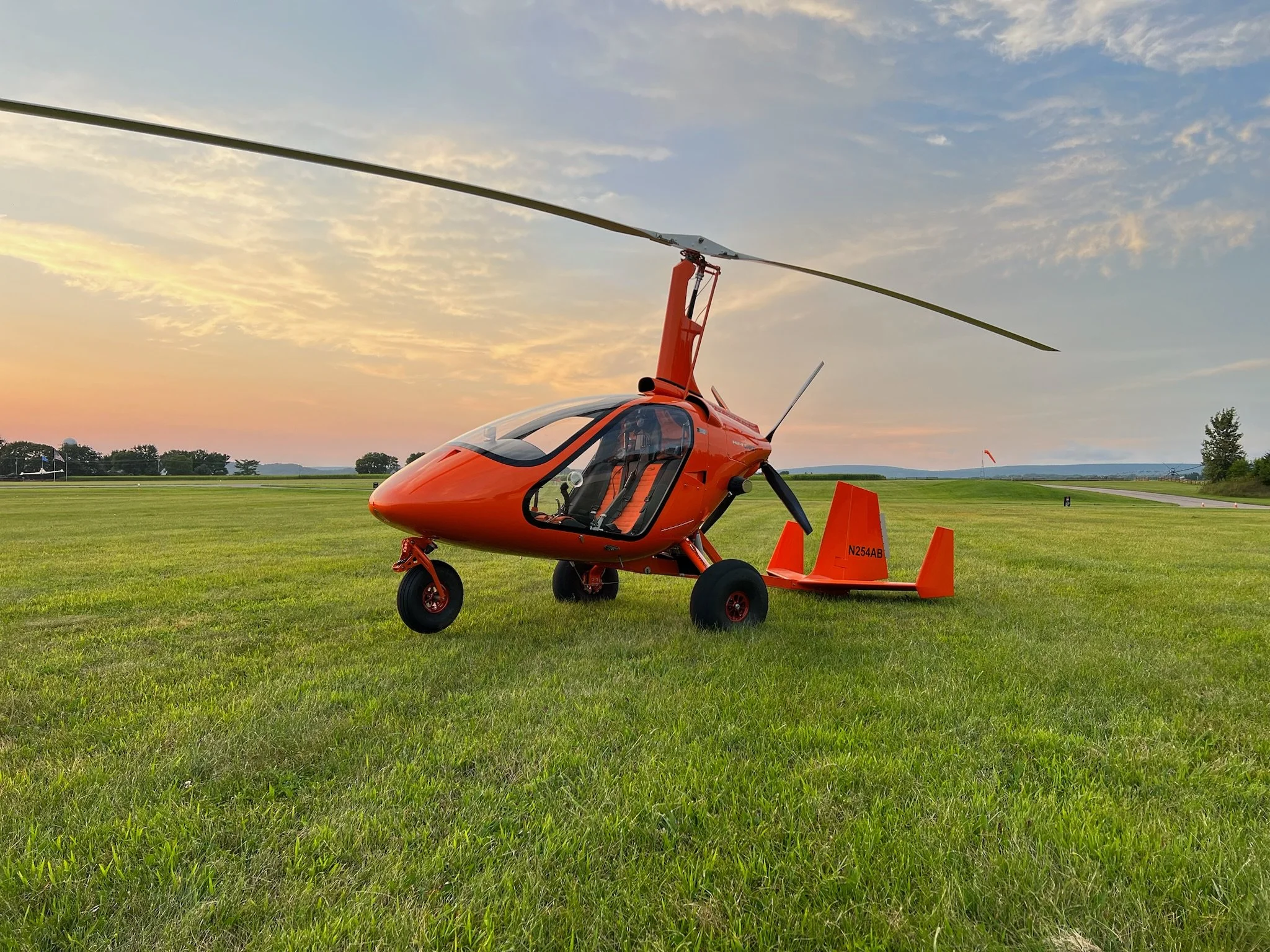 An orange M24 gyroplane is parked on a grassy field during sunset, with the sky partly cloudy and trees in the distance.