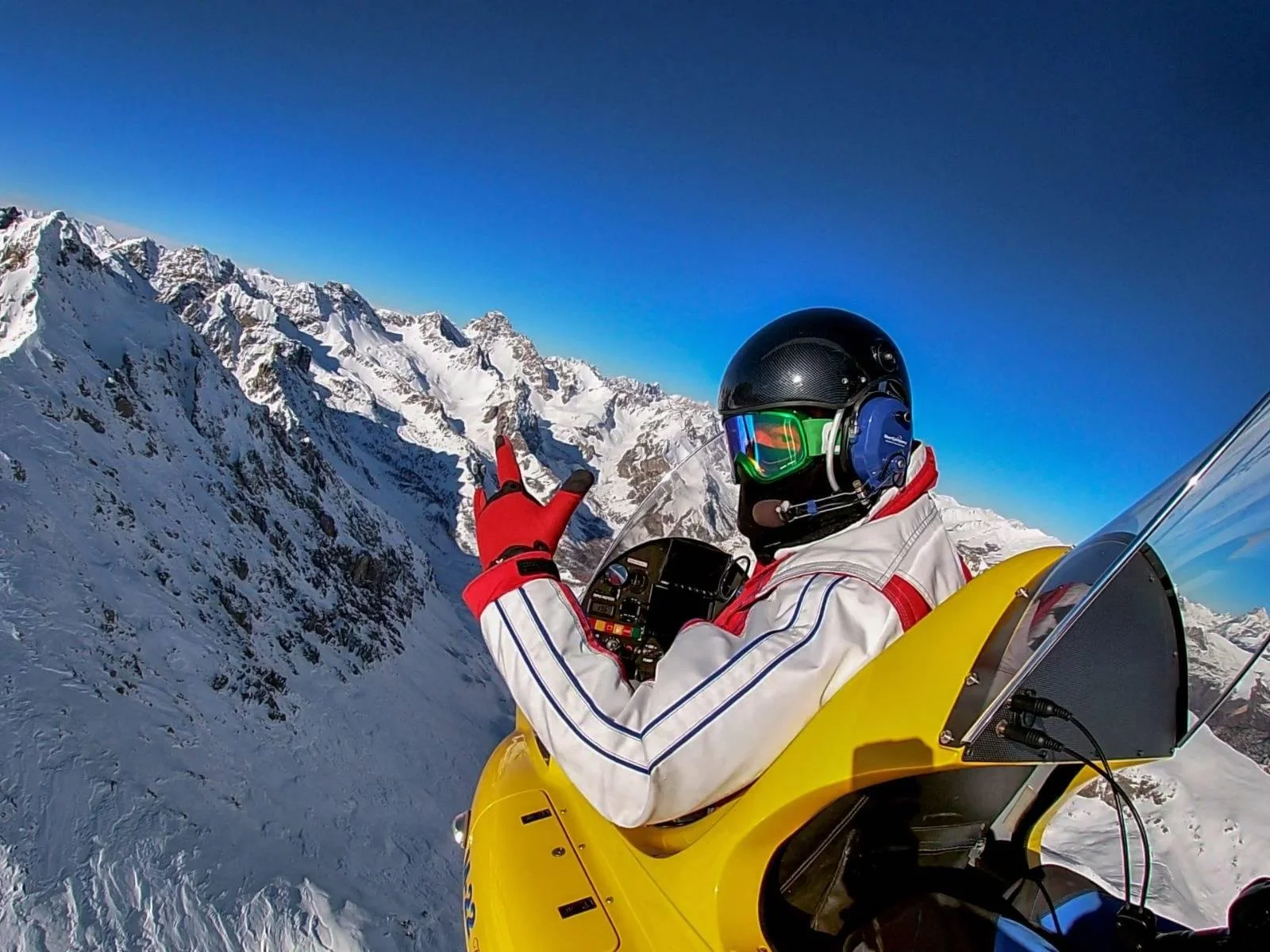 A person wearing a helmet, goggles, and a white jacket with red and blue accents, flying a yellow M22 gyroplane