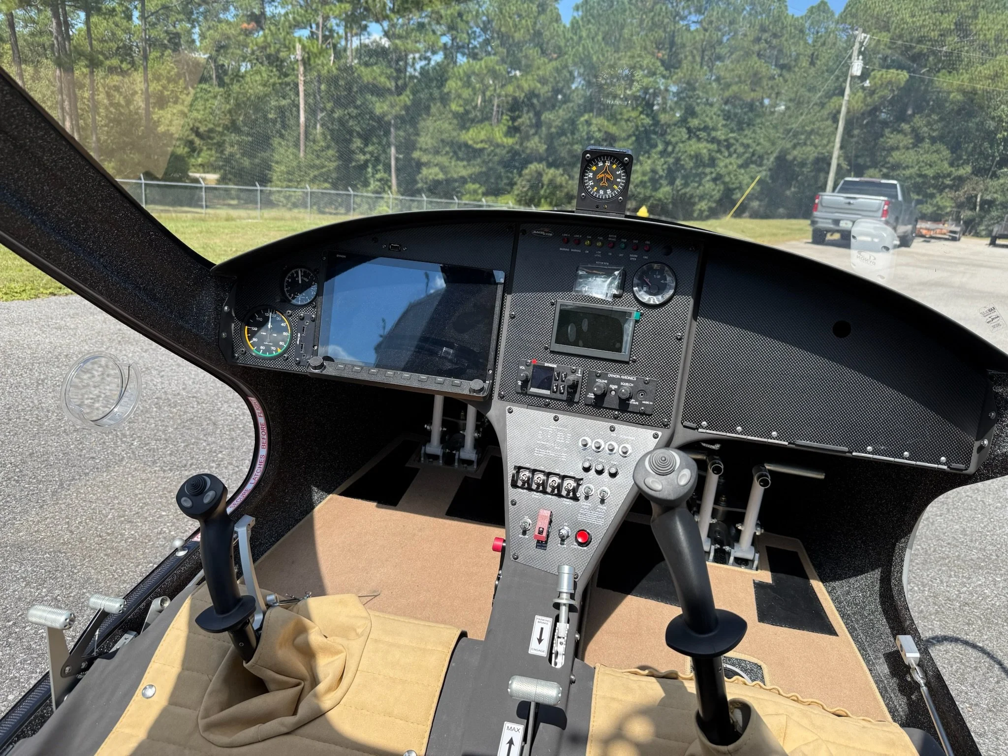 Interior view of a gyroplane cockpit with various instruments, controls, and two control sticks, with an outside scene of a parking area and trees.
