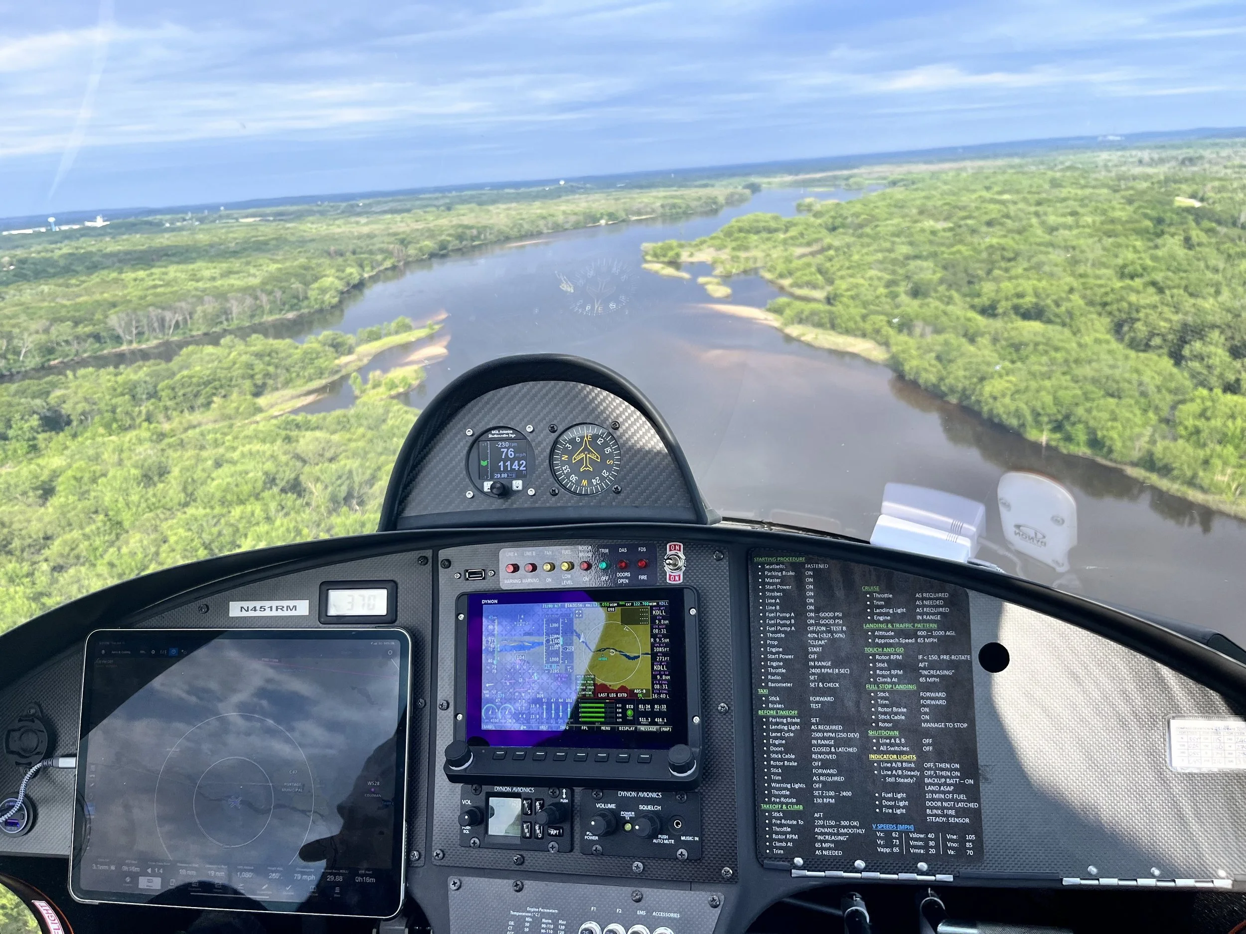 A view from inside a gyroplane cockpit, showing various flight instruments and a landscape of a winding river and lush green forests below.