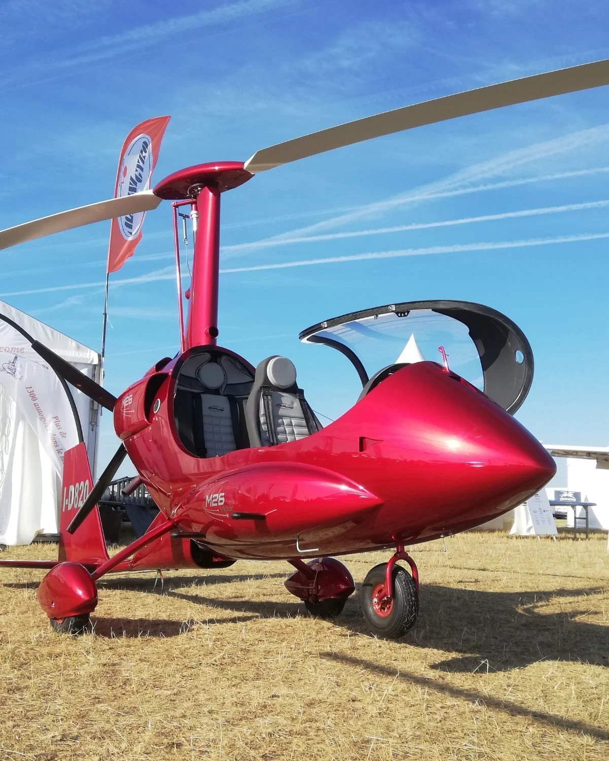 Red M26 gyroplane with bubble canopy on a grassy field under a blue sky.
