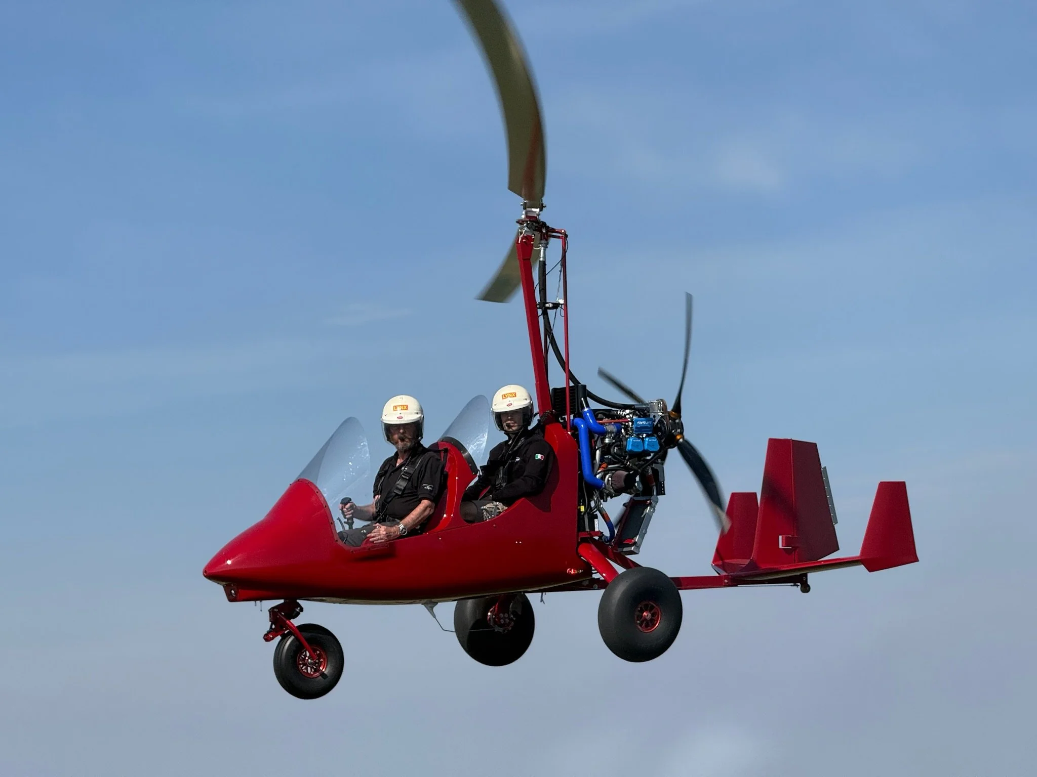 Red gyroplane with two pilots wearing white helmets flying against blue sky.