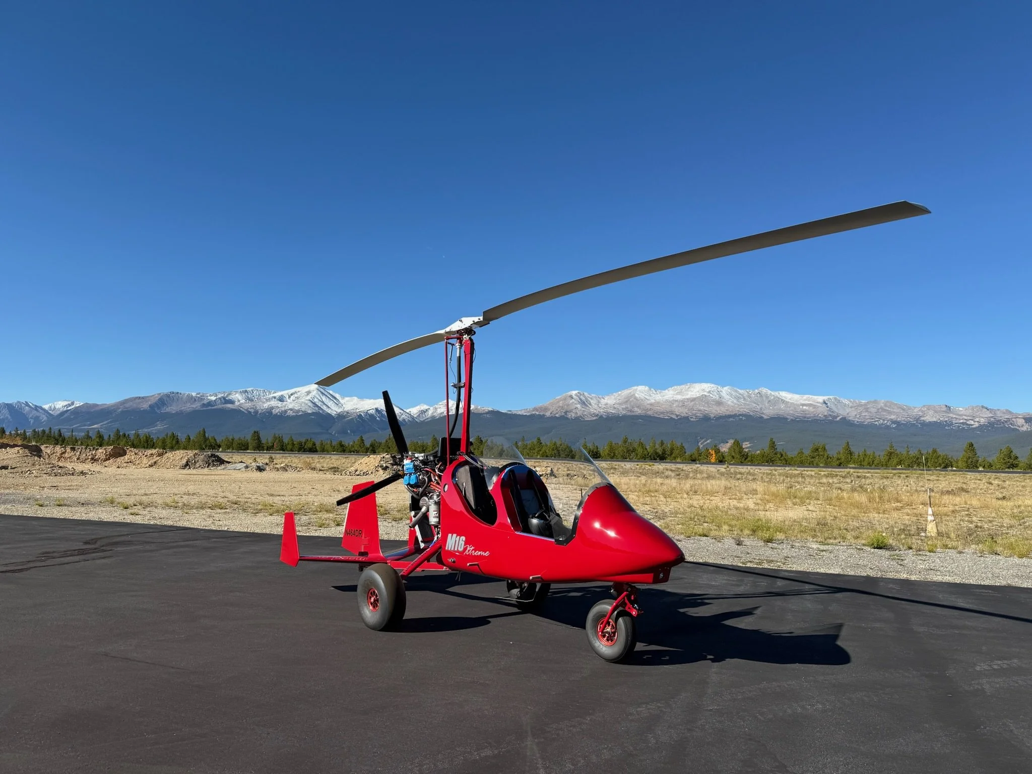 Red Gyroplane on tarmac, mountains with snow in background, clear blue sky