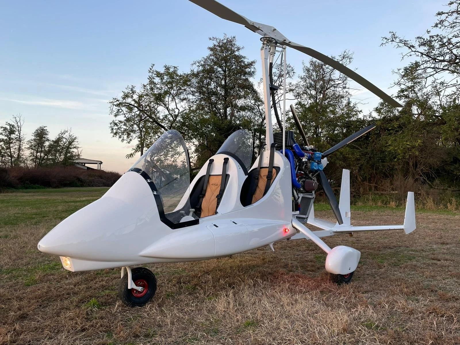 Small white M22 gyroplane parked on grassy terrain with trees in the background.