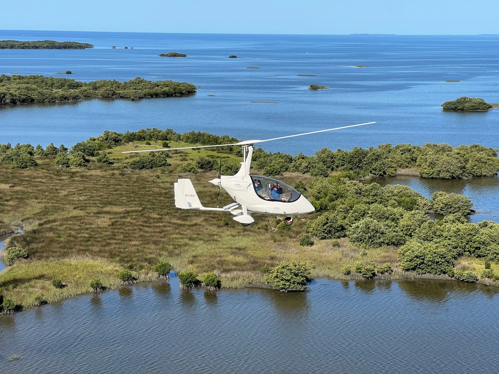 A white gyroplane flying over a lush green wetland with small bodies of water, surrounded by trees, with a large body of water with small islands in the background.