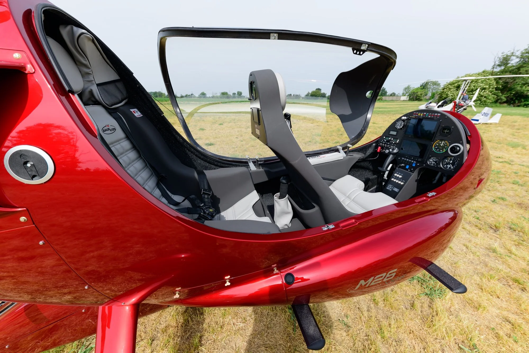 Interior of a red M26 gyroplane and advanced cockpit instruments, parked on a grassy field with other small planes and trees in the background.