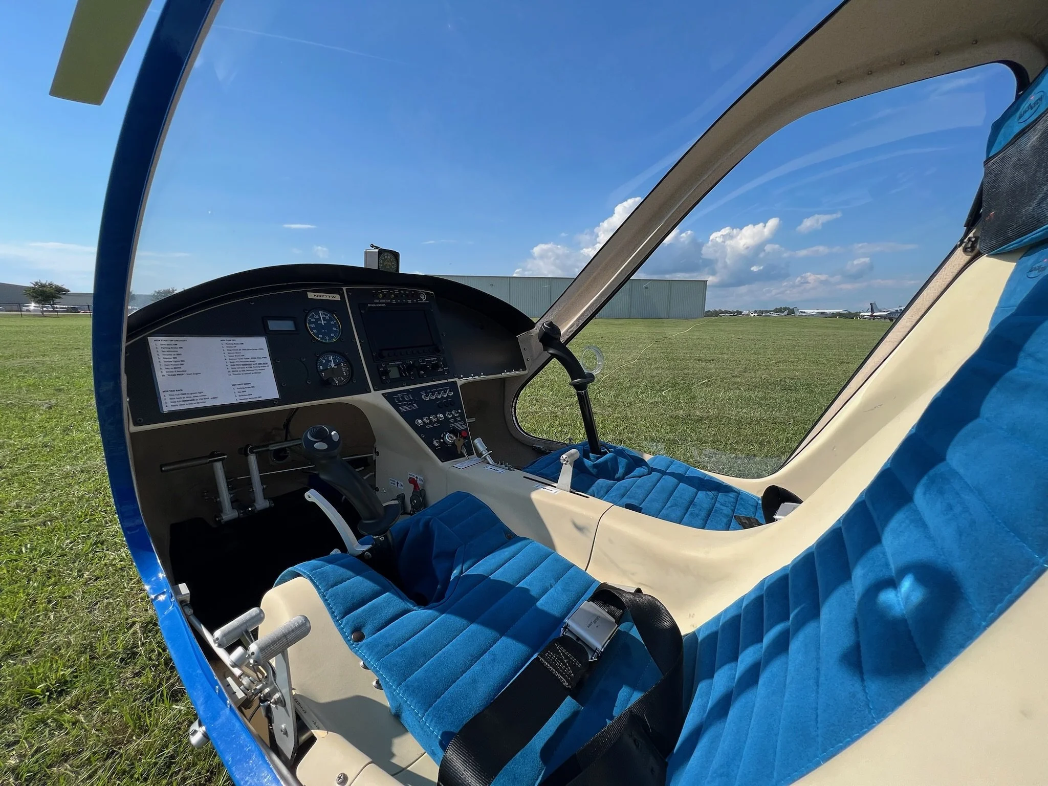 Inside view of a gyroplane cockpit on a grassy airfield, showing two blue padded seats, flight instruments, and a clear sky with a few clouds.