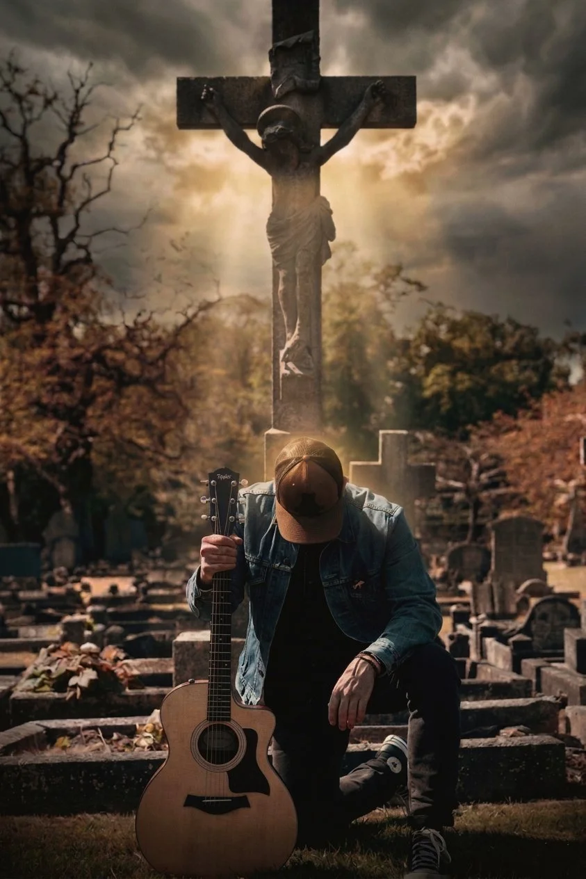 A man kneeling in a cemetery holding a guitar, with a large crucifix statue in the background and cloudy skies overhead.