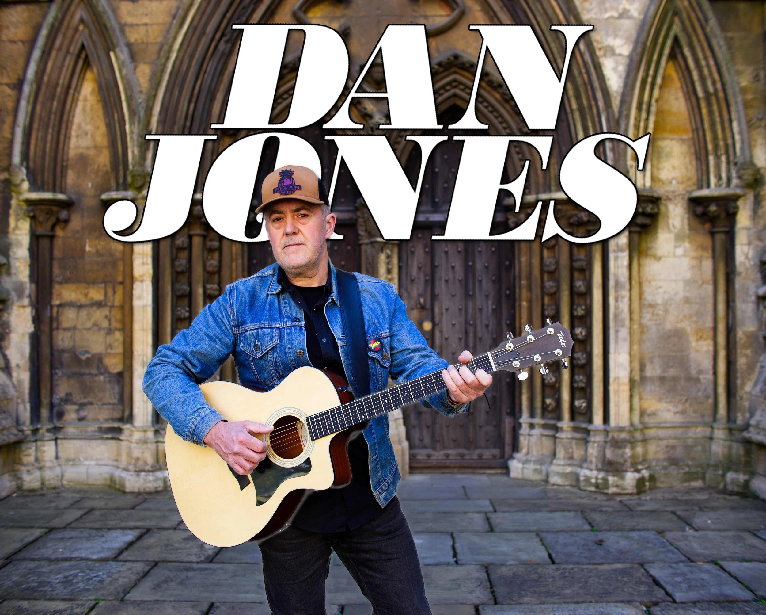 A man wearing a cap, denim jacket, and black shirt playing an acoustic guitar in front of an old church door.