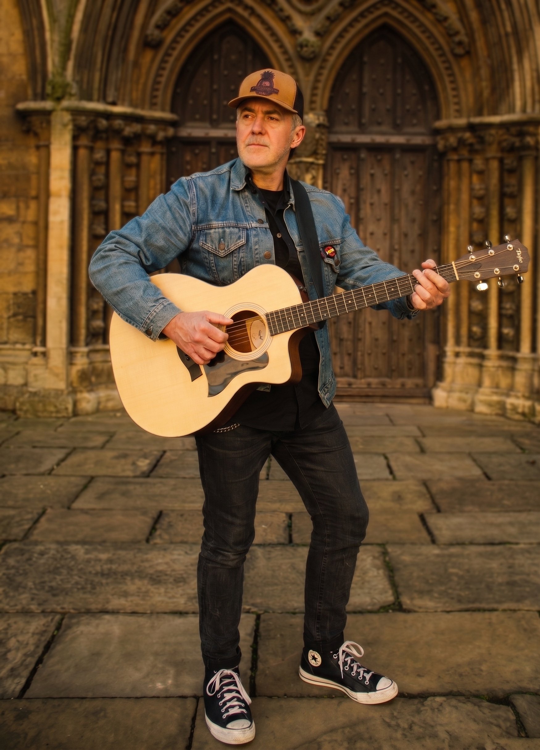 A man wearing a tan and black trucker cap, denim jacket, and black jeans is playing an acoustic guitar on a cobblestone street in front of a large wooden door with decorative stone archway.