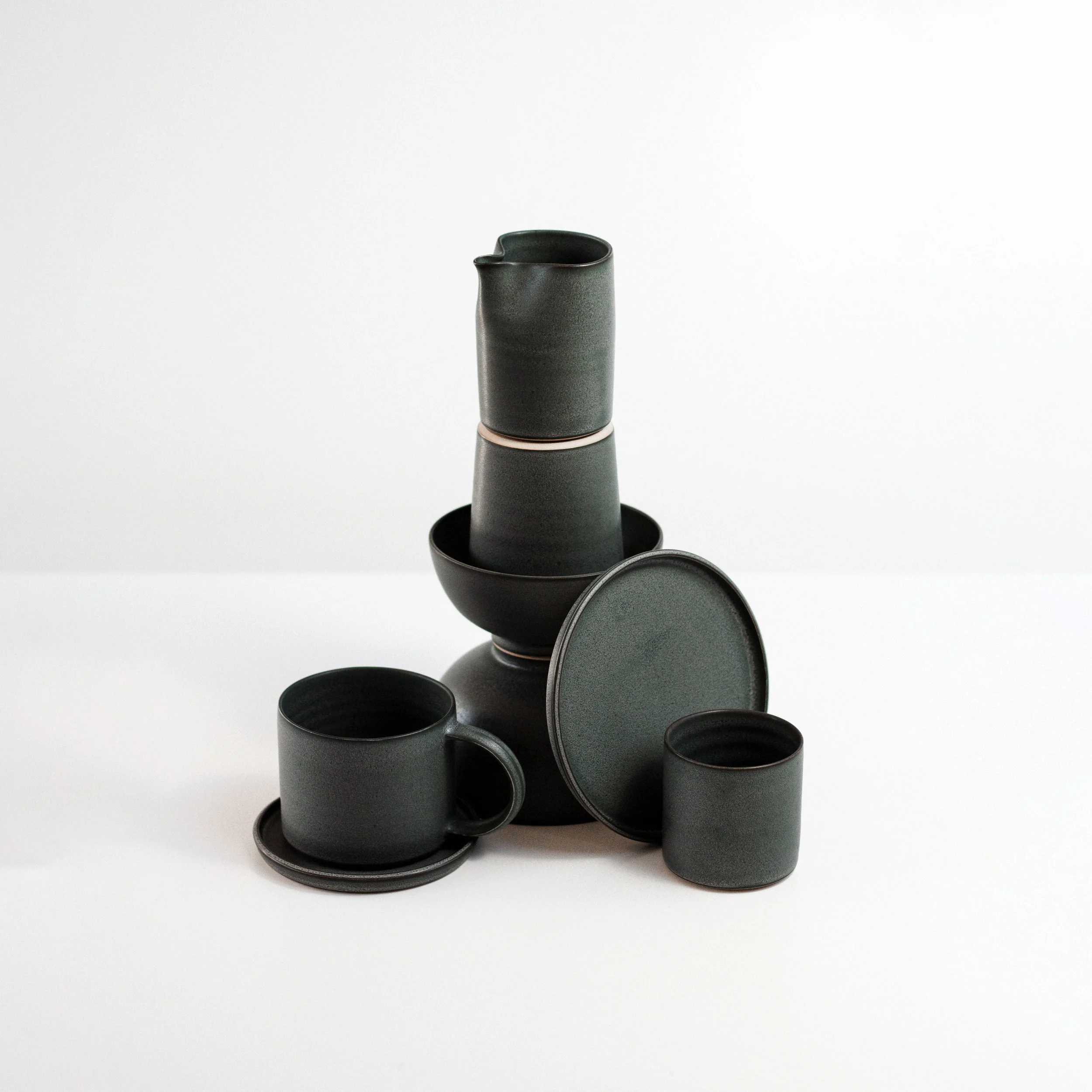 Black ceramic dinnerware, including a mug, a small cup, a bowl, and plates, arranged on a white surface with a plain white background.
