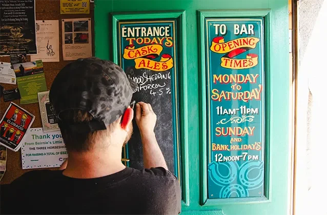 A man in a cap writing the opening times of the Three Horseshoes pub in Bradford on Avon on a chalk board on the front door