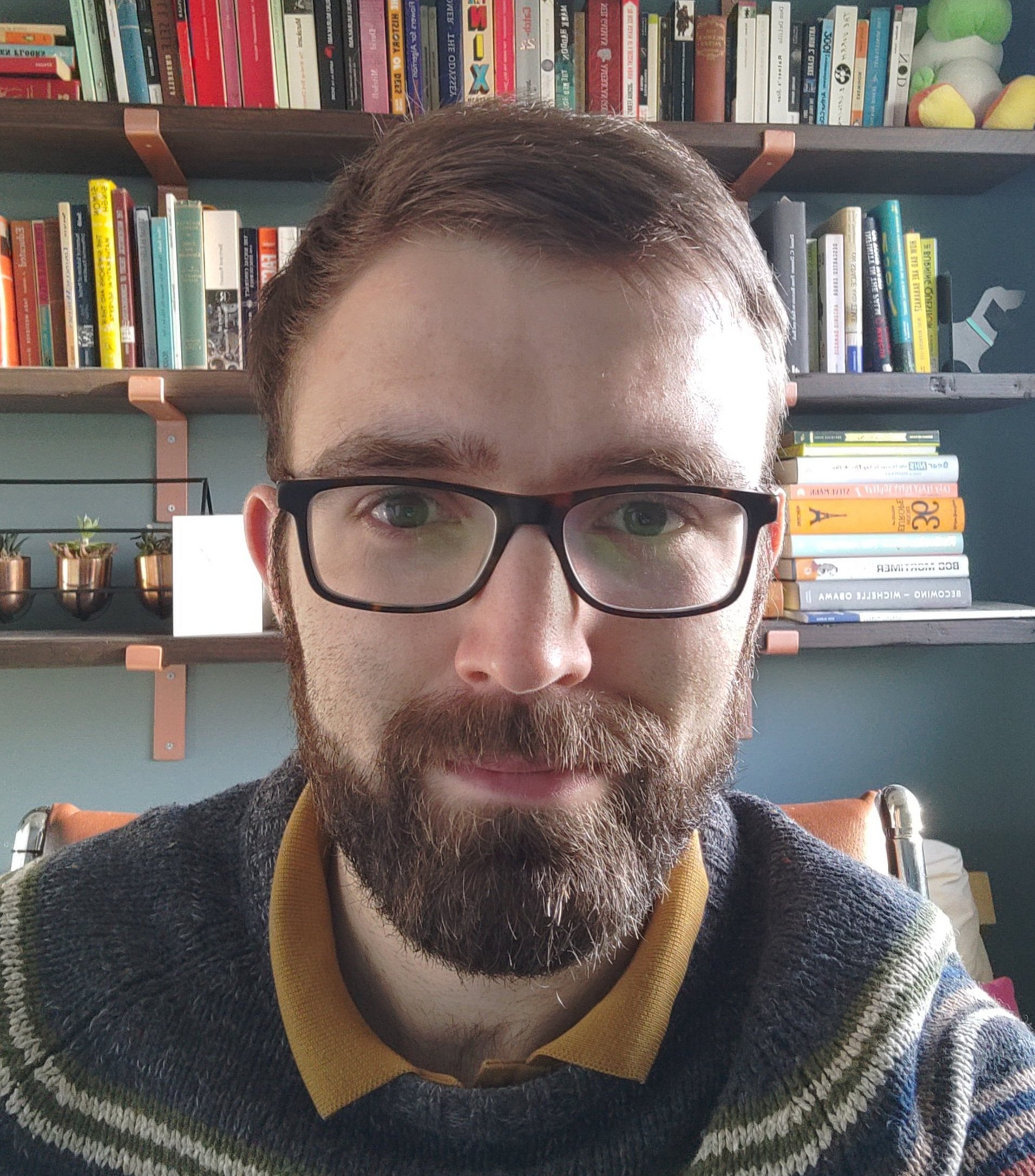 Headshot of Carl Whitfield, a man with short brown hair, glasses and a beard, sat in front of some shelves of books.