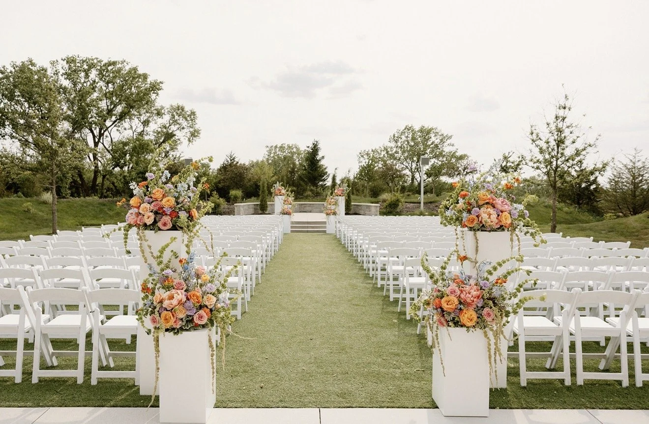 Outdoor wedding ceremony setup with white folding chairs and colorful floral arrangements lining a grassy aisle