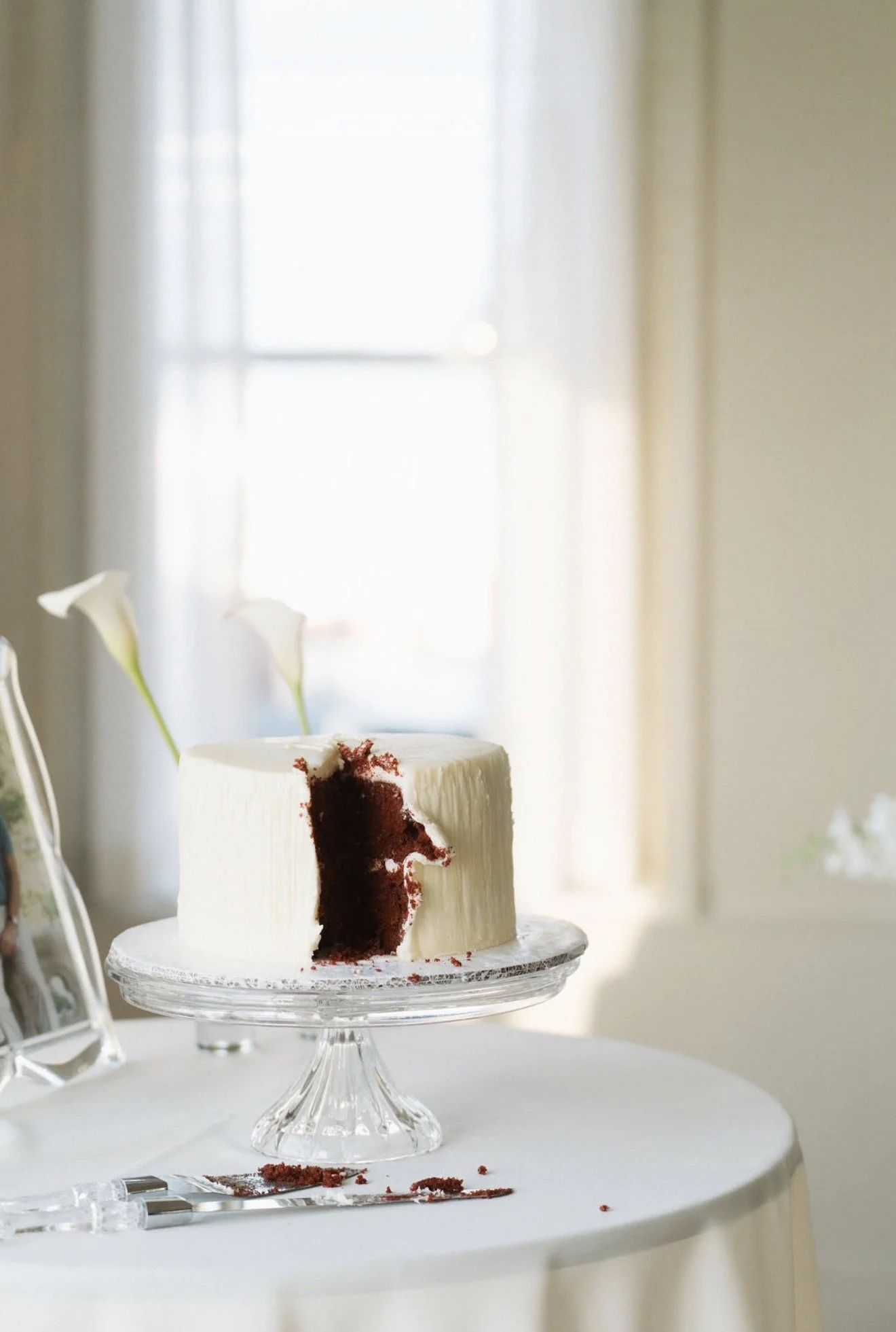 White-frosted red velvet cake with a slice removed, displayed on a glass cake stand beside serving utensils and white calla lilies.