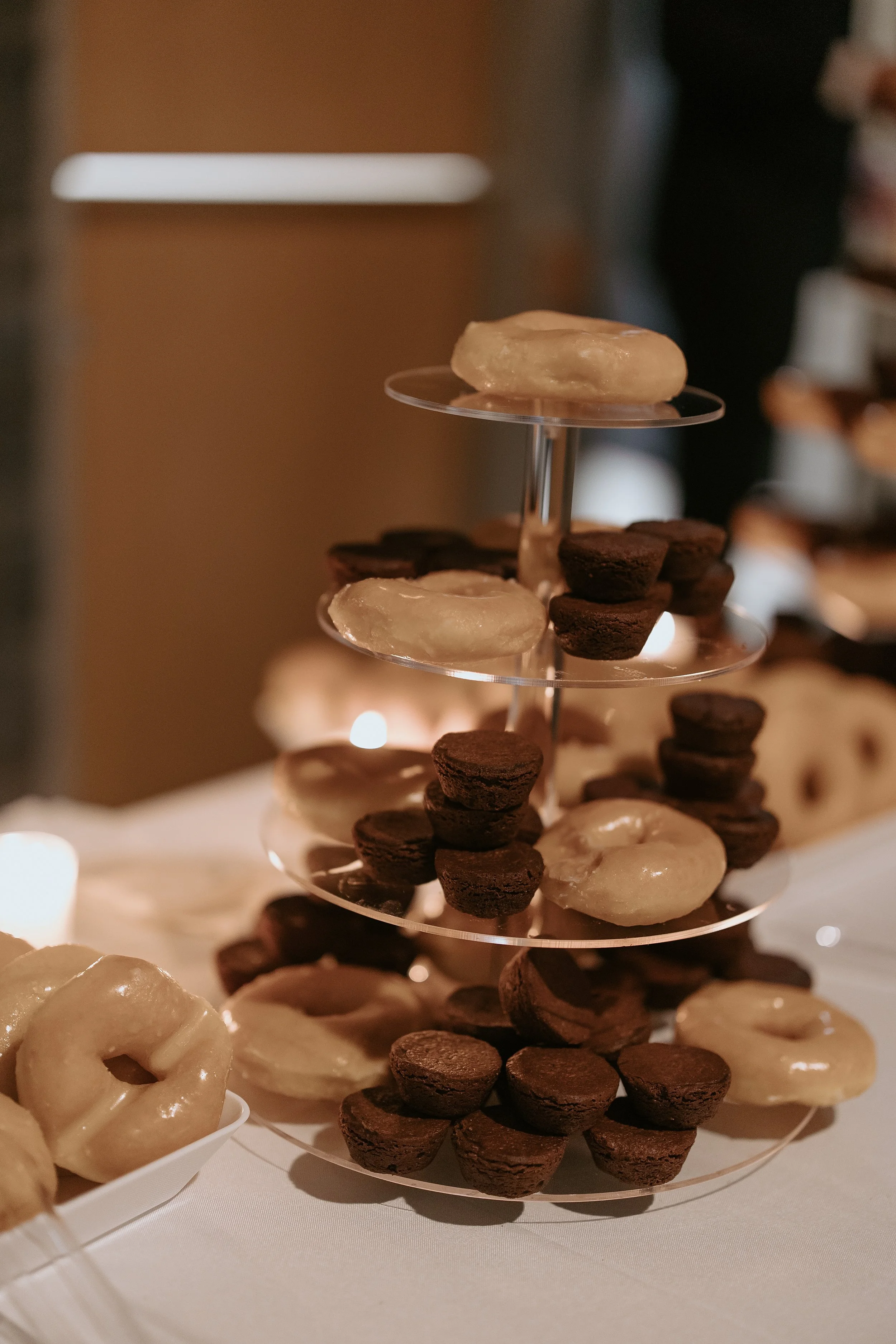 Tiered glass dessert stand displaying chocolate and glazed mini donuts at a warmly lit dessert table