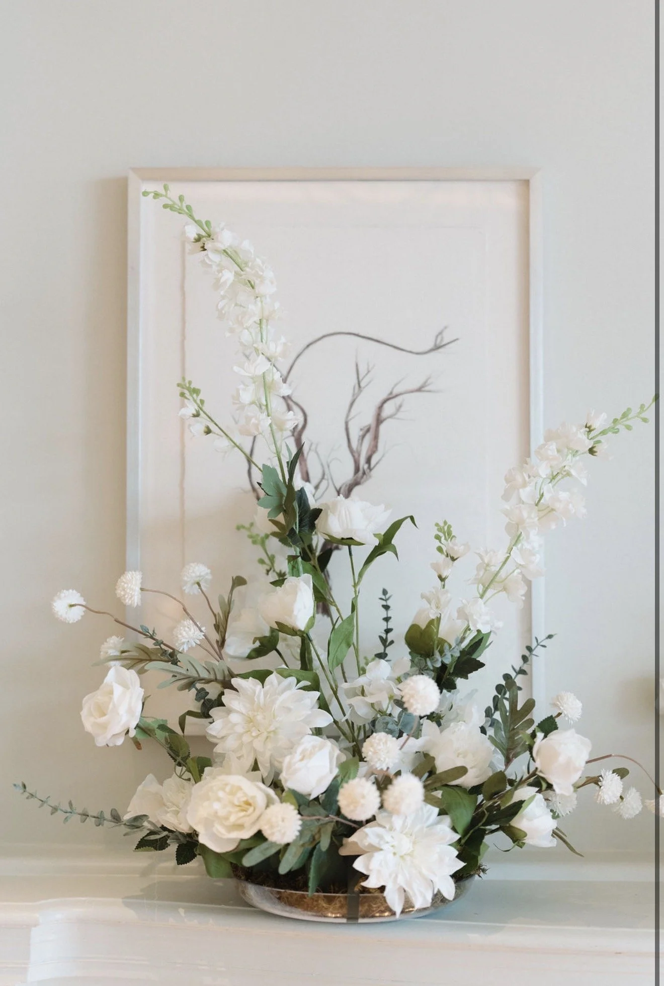 Large white floral arrangement with greenery in a shallow bowl, displayed on a mantel beneath framed botanical artwork