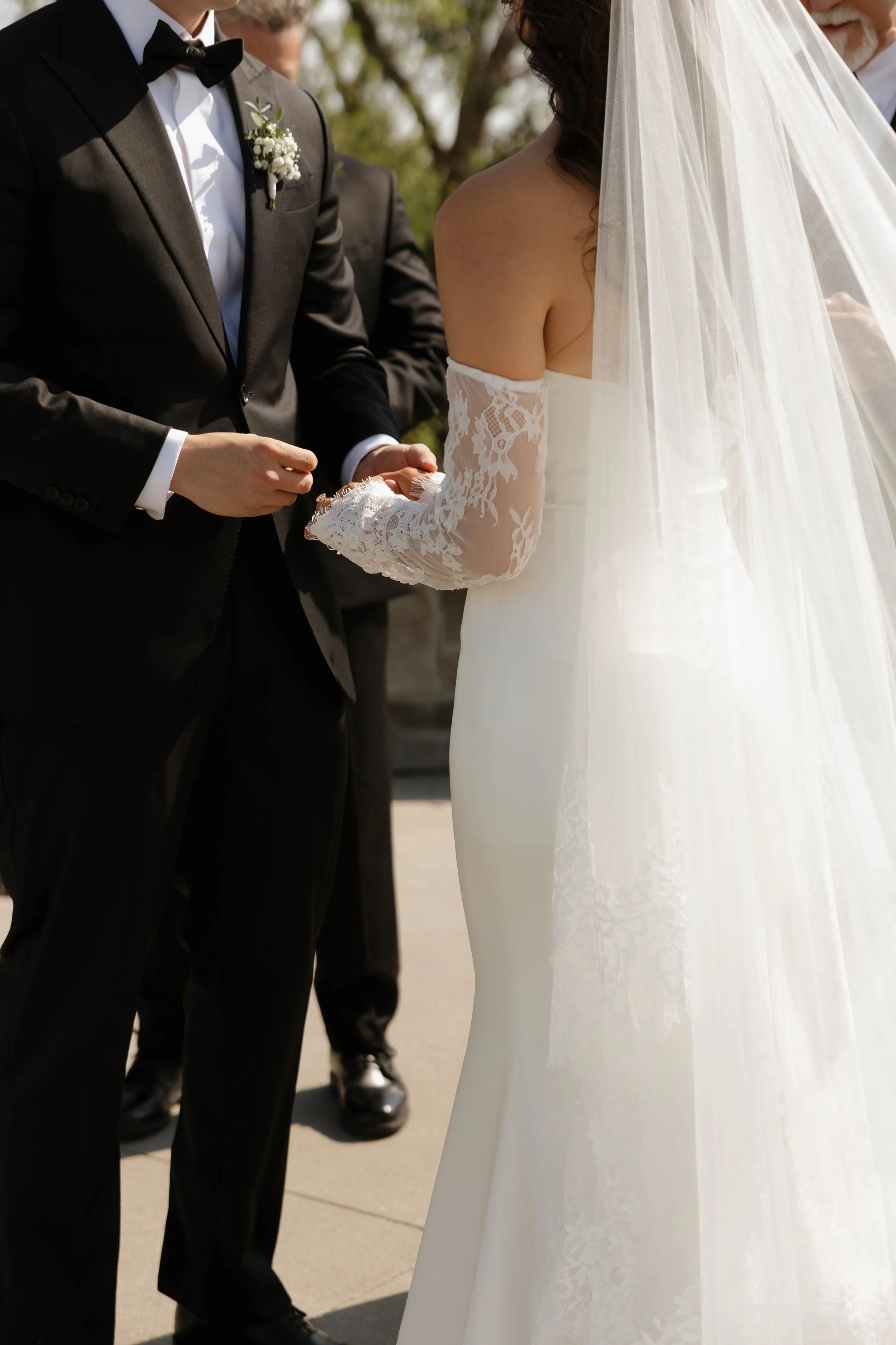 Bride in a lace wedding dress and veil standing beside a groom in a black tuxedo with a boutonniere during an outdoor wedding ceremony