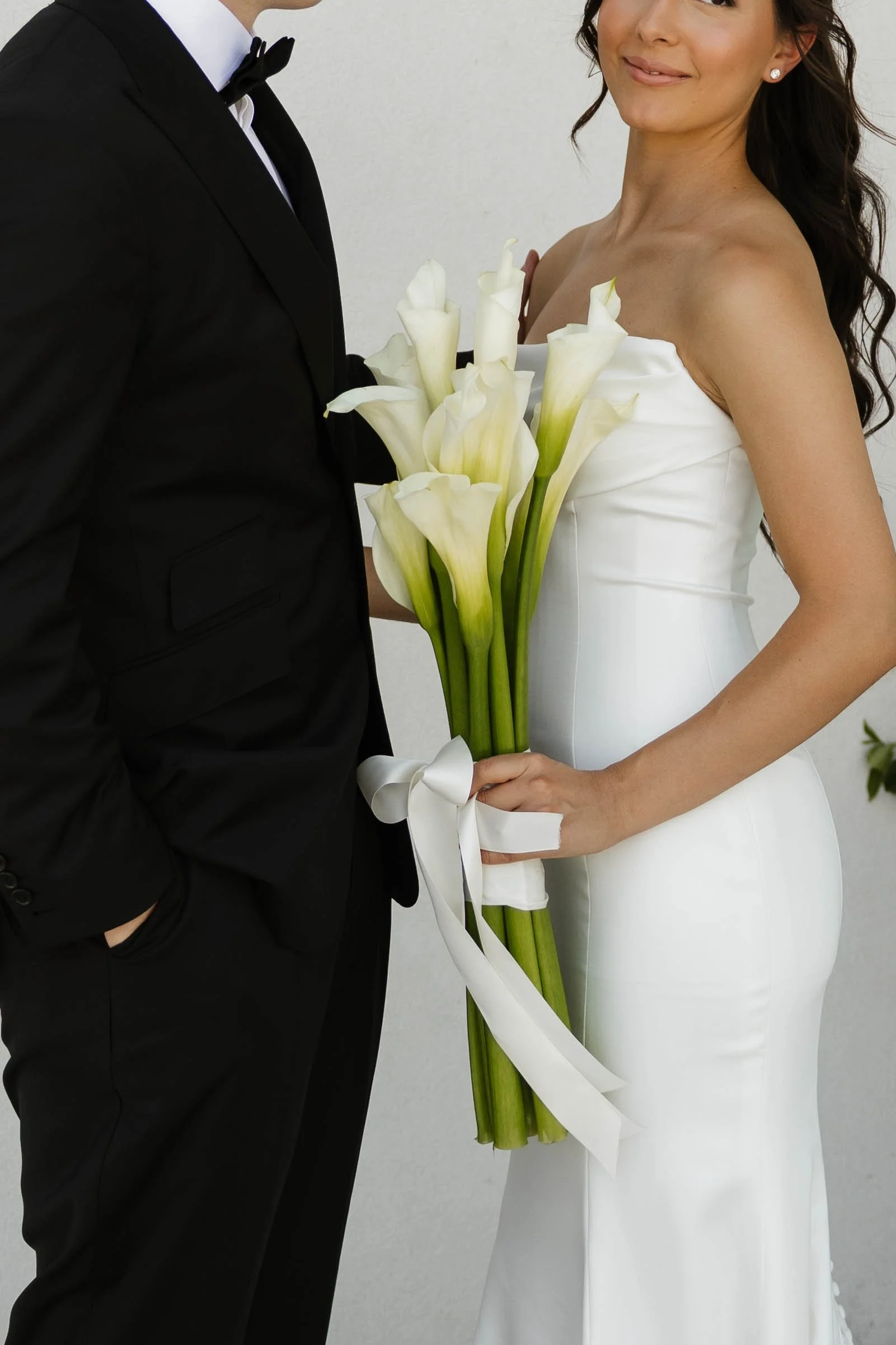 Bride in a white strapless wedding dress holding a bouquet of white calla lilies beside a groom in a black tuxedo