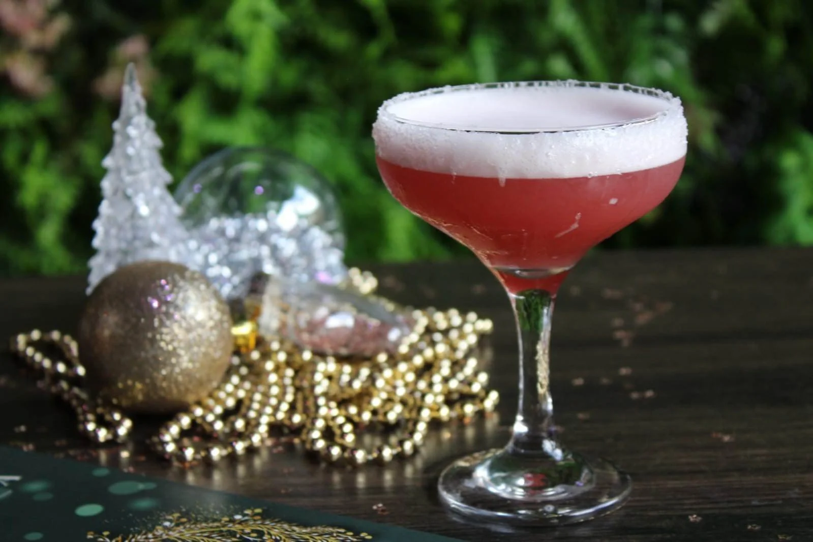 A pink cocktail in a coupe glass with a salt rim, surrounded by holiday decorations including gold and silver ornaments, beads, and small decorative trees, on a dark wooden surface with a green leafy background.