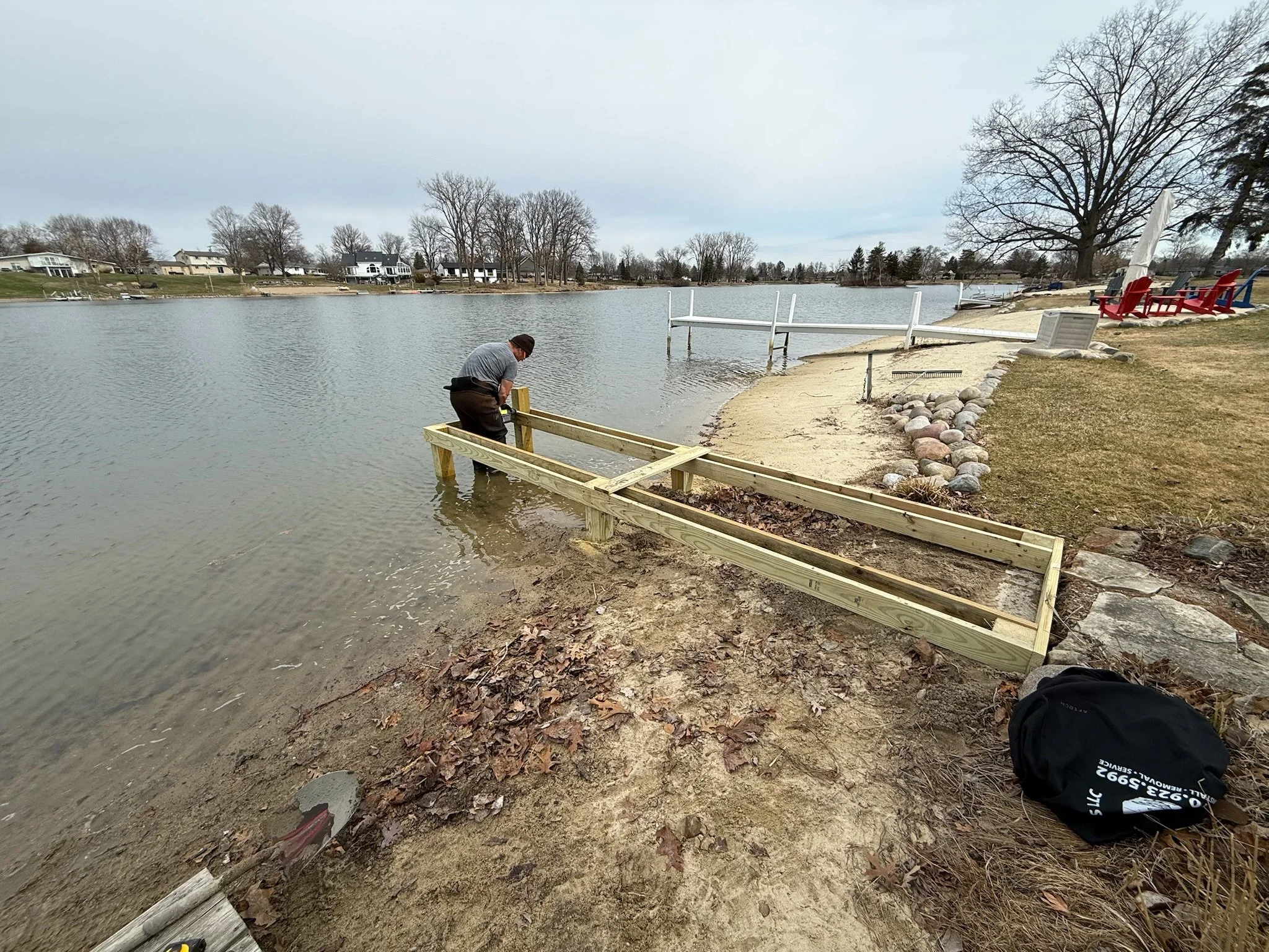 Man installing a wooden dock by a lake on a cloudy day, with houses in the background, and Adirondack chairs on the shore.
