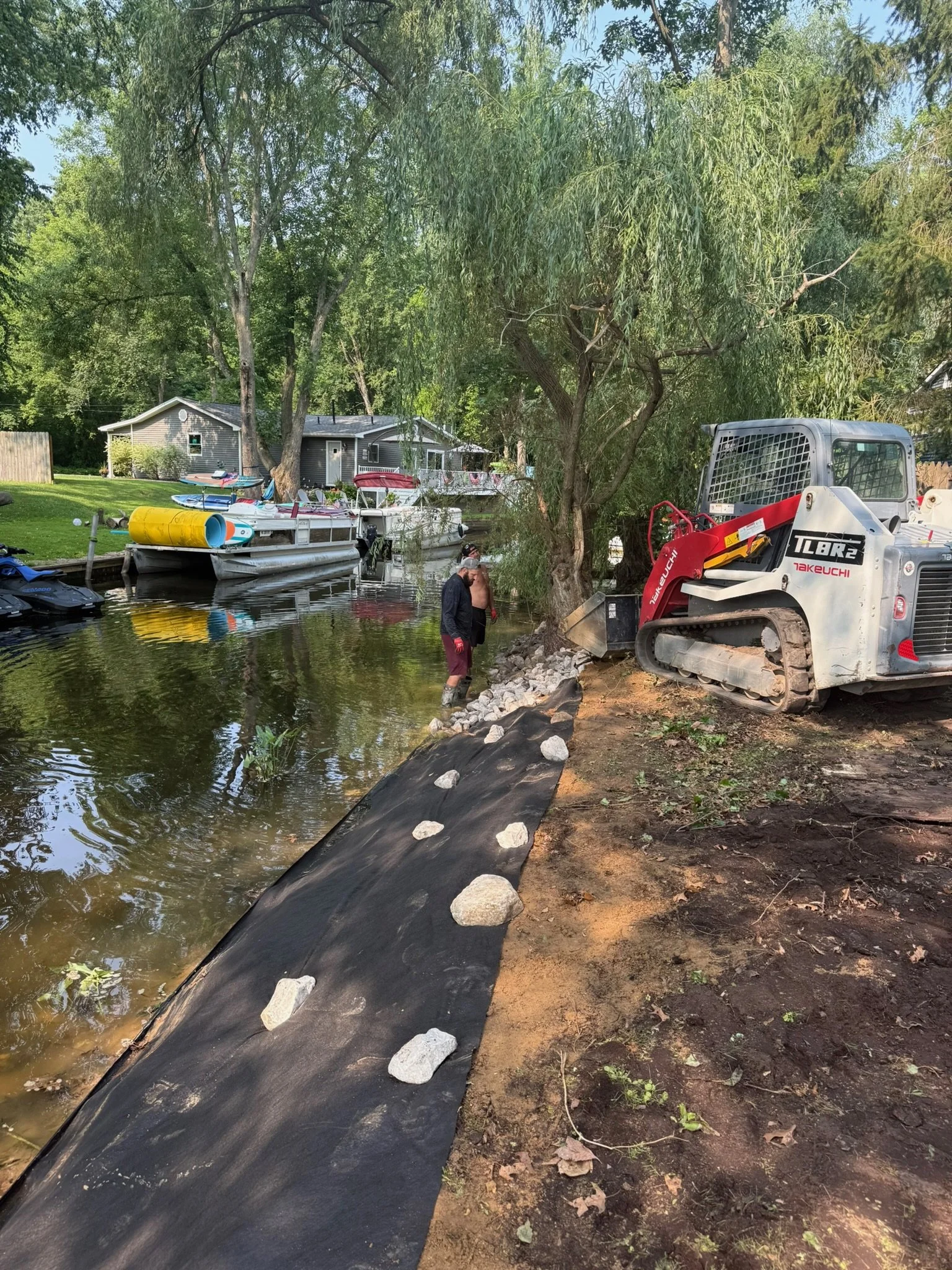 A backyard canal with boats and a construction machine on the bank, surrounded by trees and a house in the background.