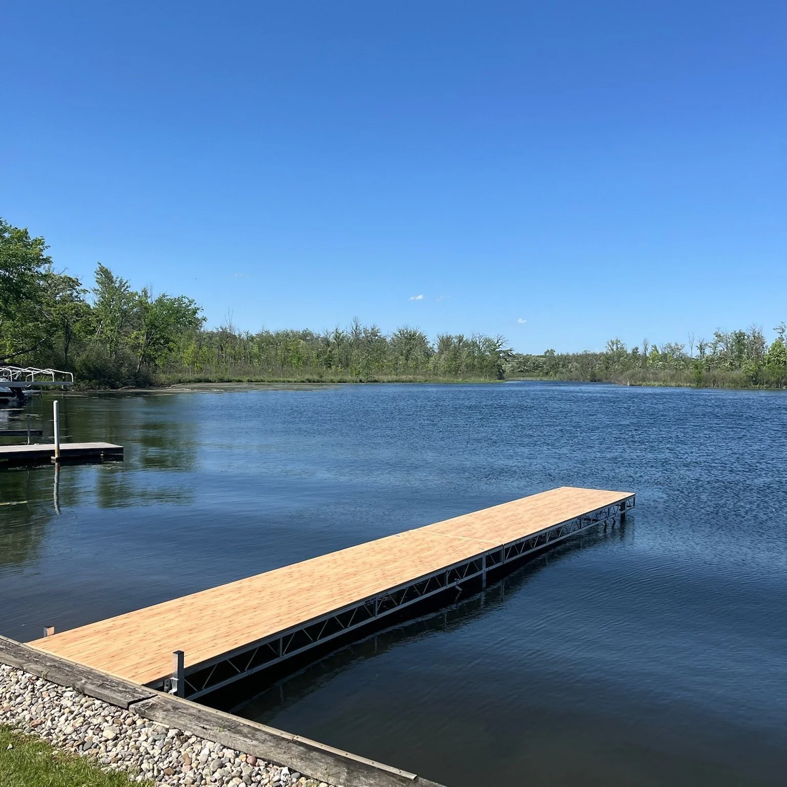 A calm lake with a small dock extending into the water, surrounded by trees under a clear blue sky.