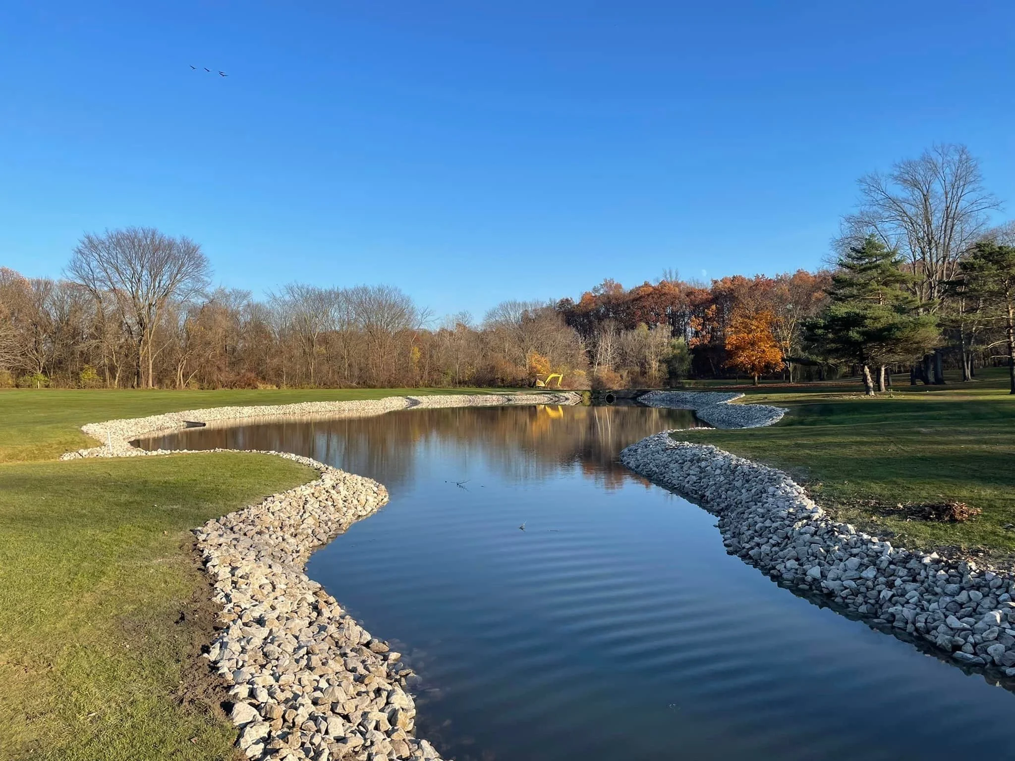 A peaceful park scene with a narrow waterway lined with rocks. The water reflects the blue sky, and in the background, there are trees with autumn foliage and some bare branches. The sky is clear, and there are three birds flying in formation.