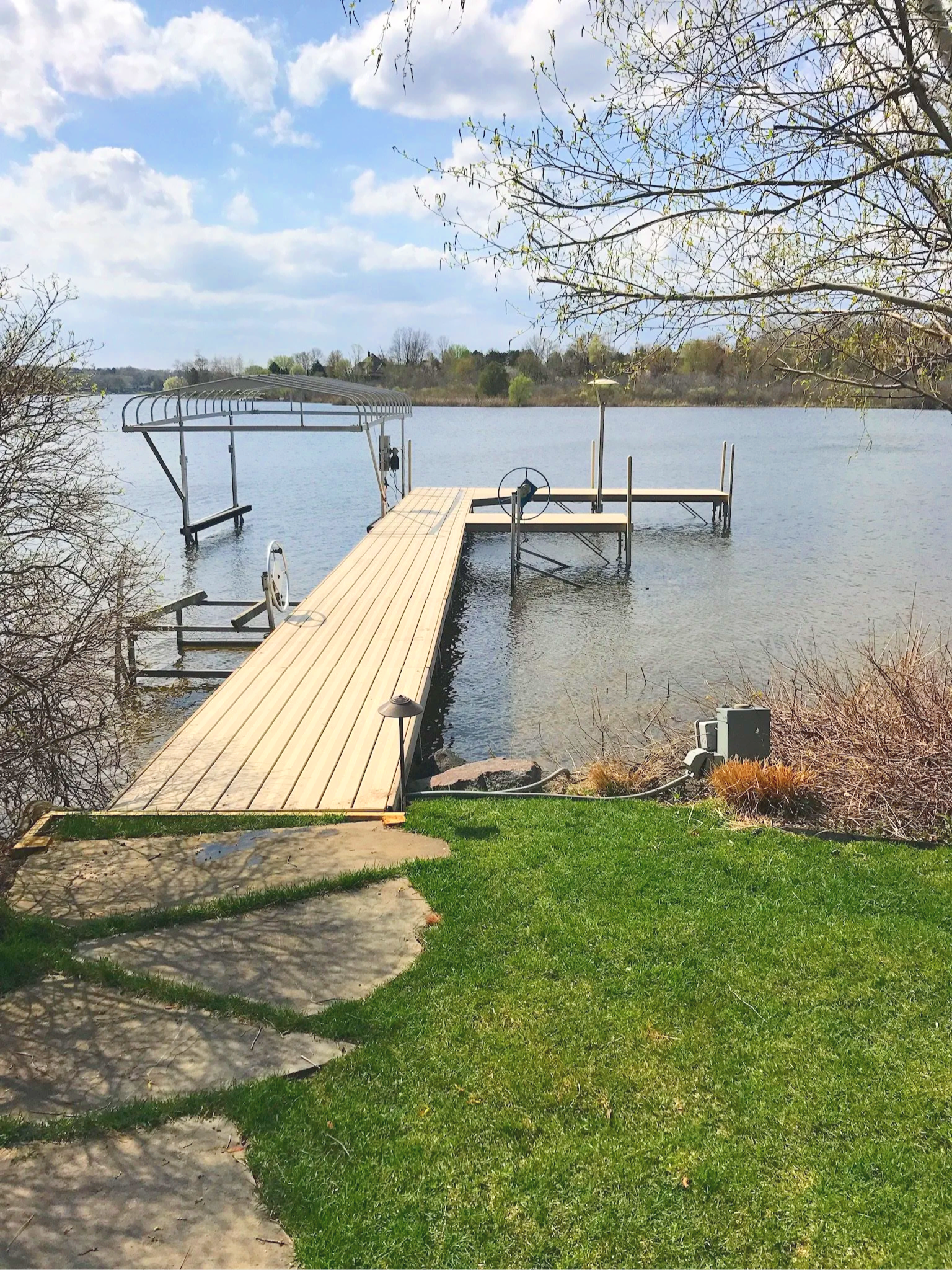 A wooden dock extends over a calm lake, with a metal boat lift on the left and a small boat lift on the right. There are leafless tree branches extending over the scene, and a grassy area with a concrete walkway in the foreground. The sky is partly cloudy.