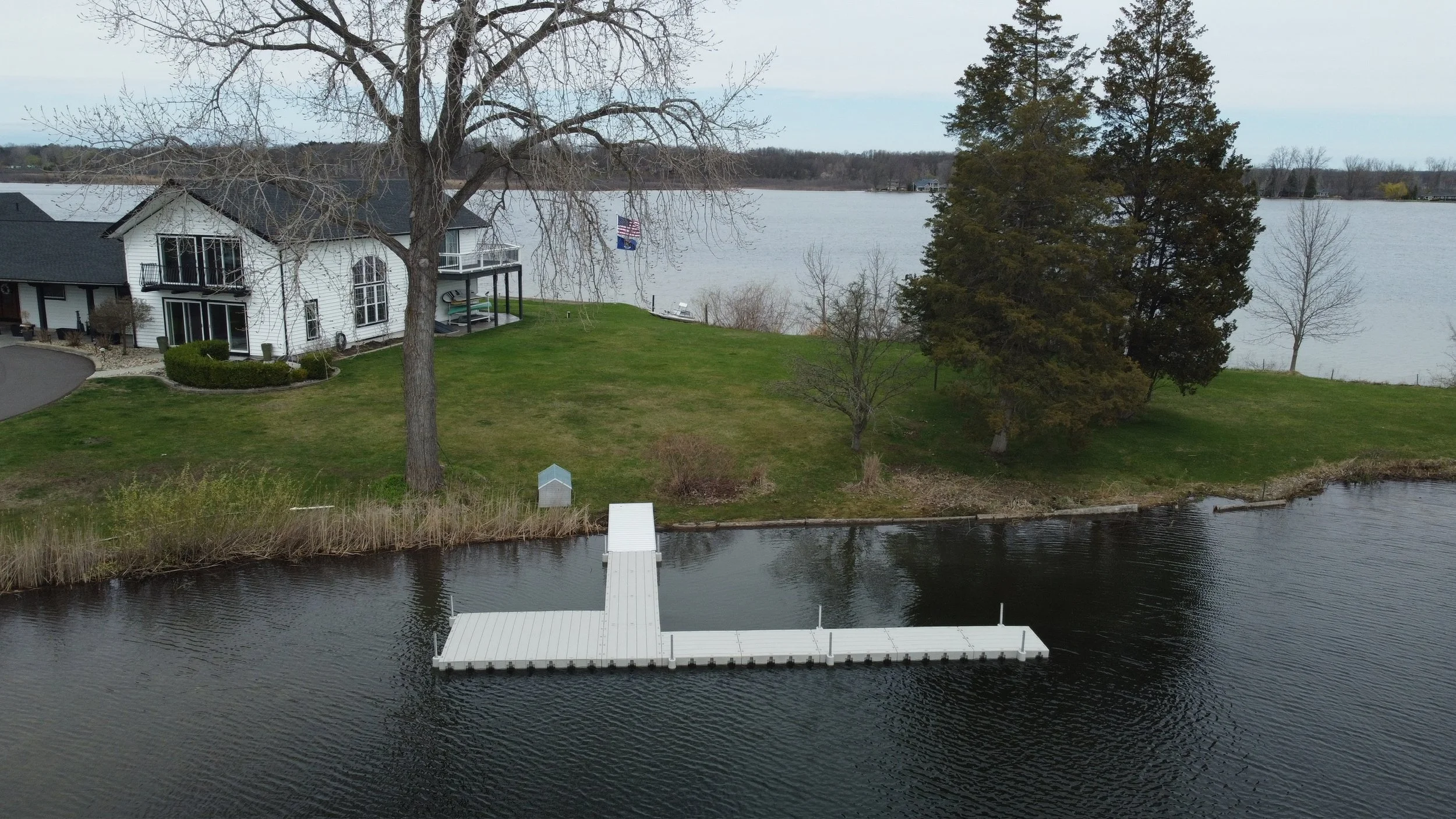A lakeside residential area with a white house, trees, grassy lawn, and a dock extending into the water.