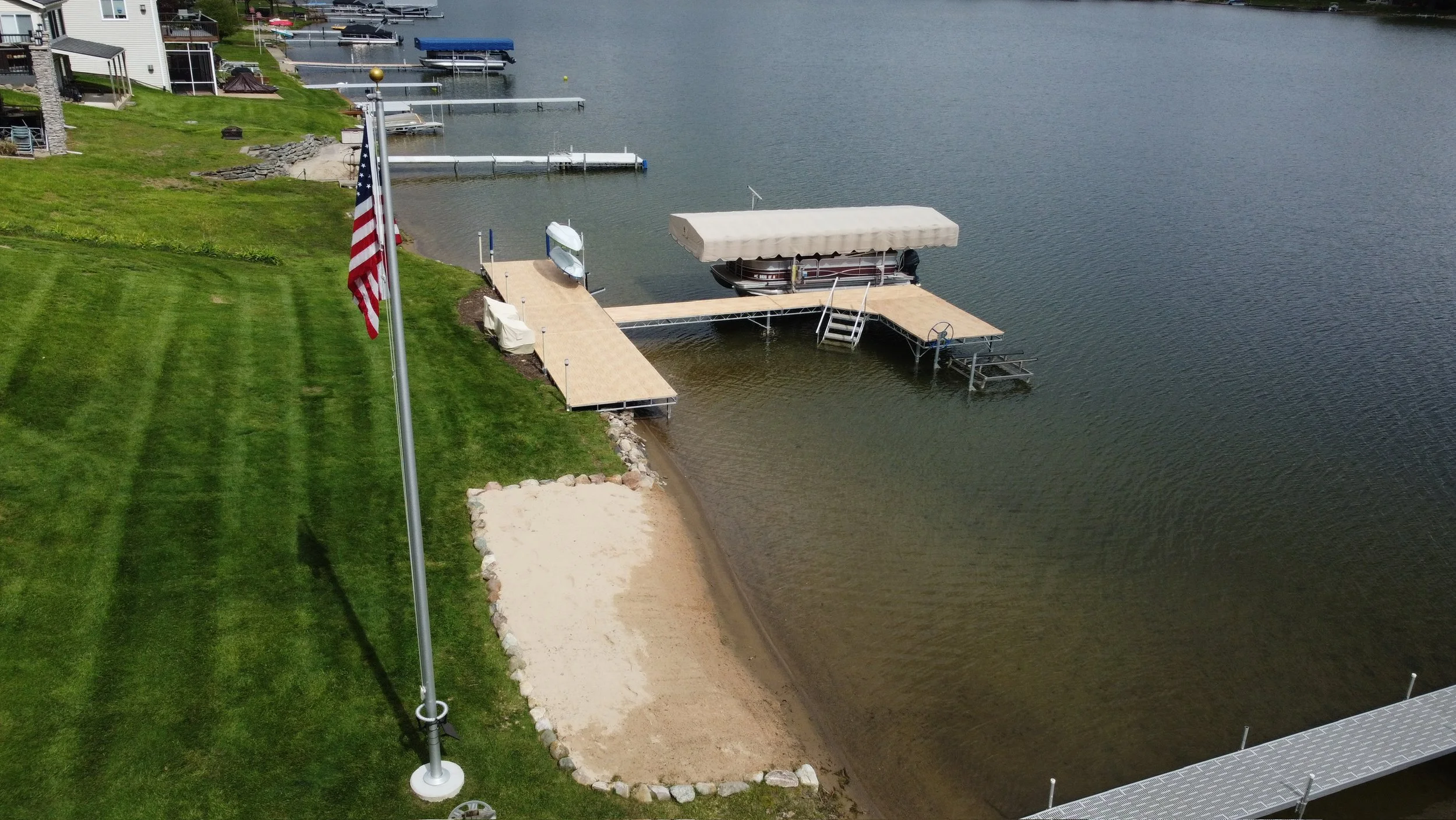 A lakeside backyard featuring a well-maintained green lawn, a flagpole with an American flag, a sandy beach area, and a dock with a boat covered by a canopy. Several small boats are moored at the docks, and there are residential houses visible in the