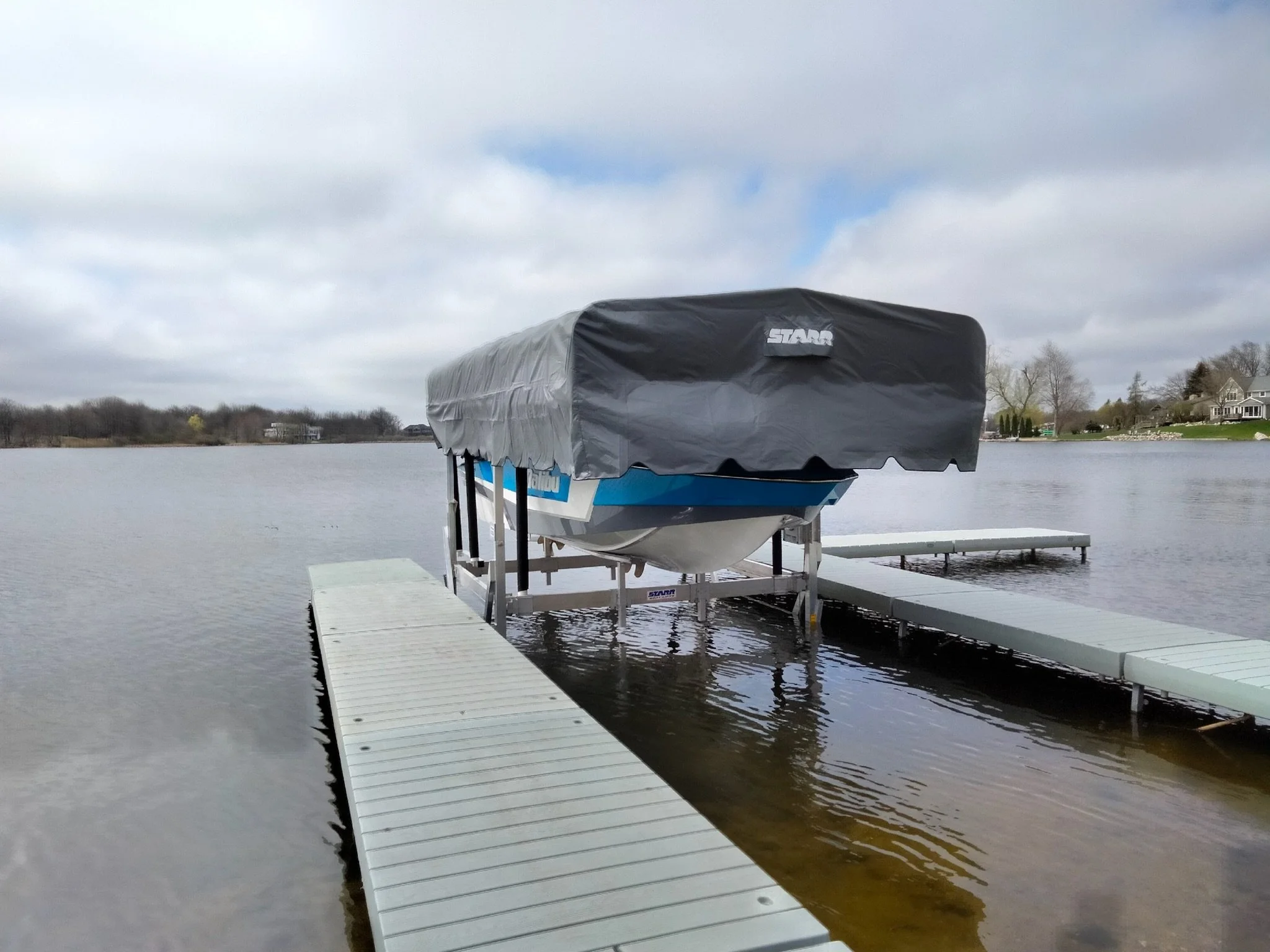A boat docked at a pier on a calm lake with a black cover over the boat, a cloudy sky, and houses in the background.