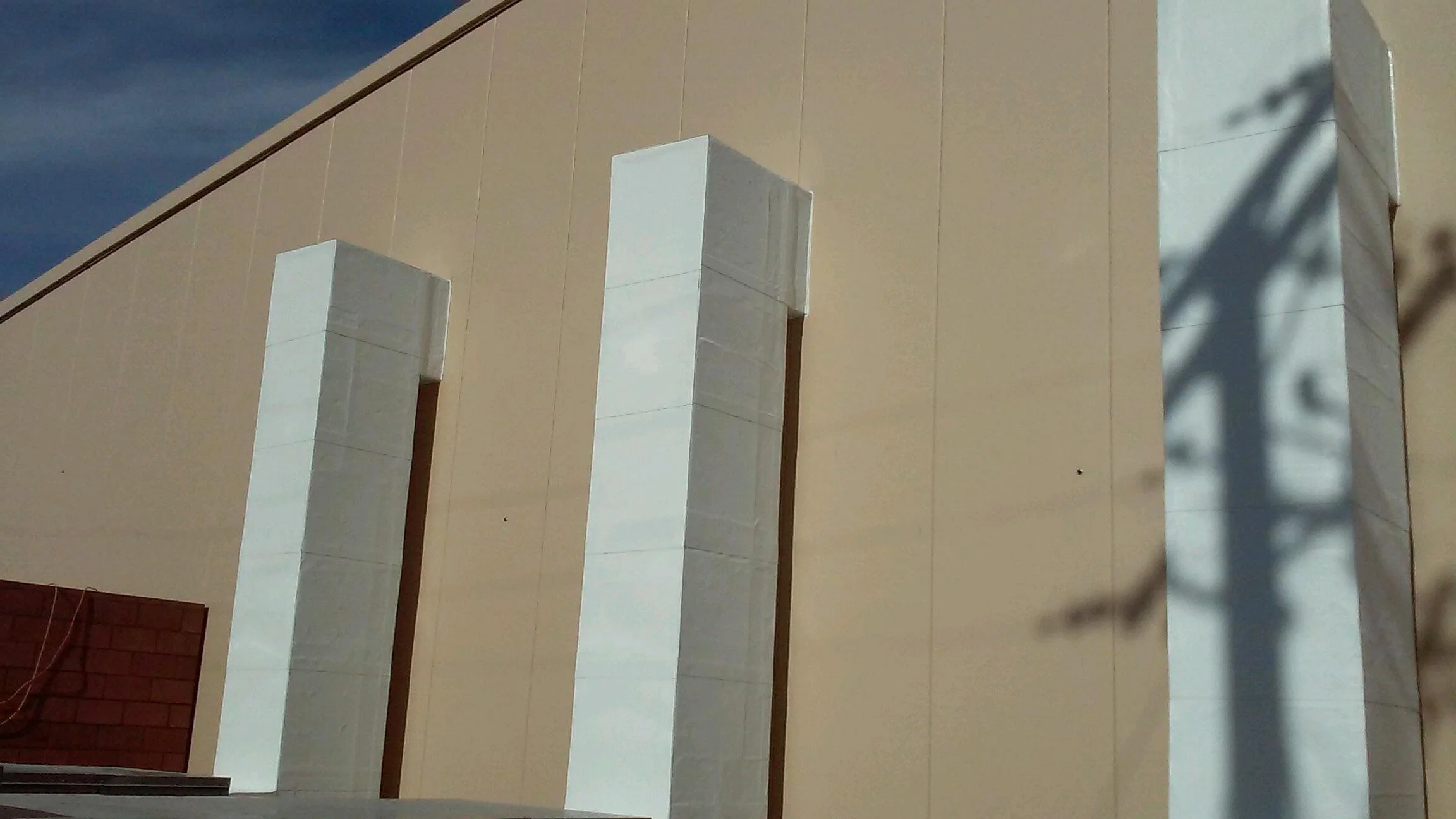 Close-up view of a modern architectural structure with beige walls, white vertical columns, and a shadow of a tree cast on the right side.