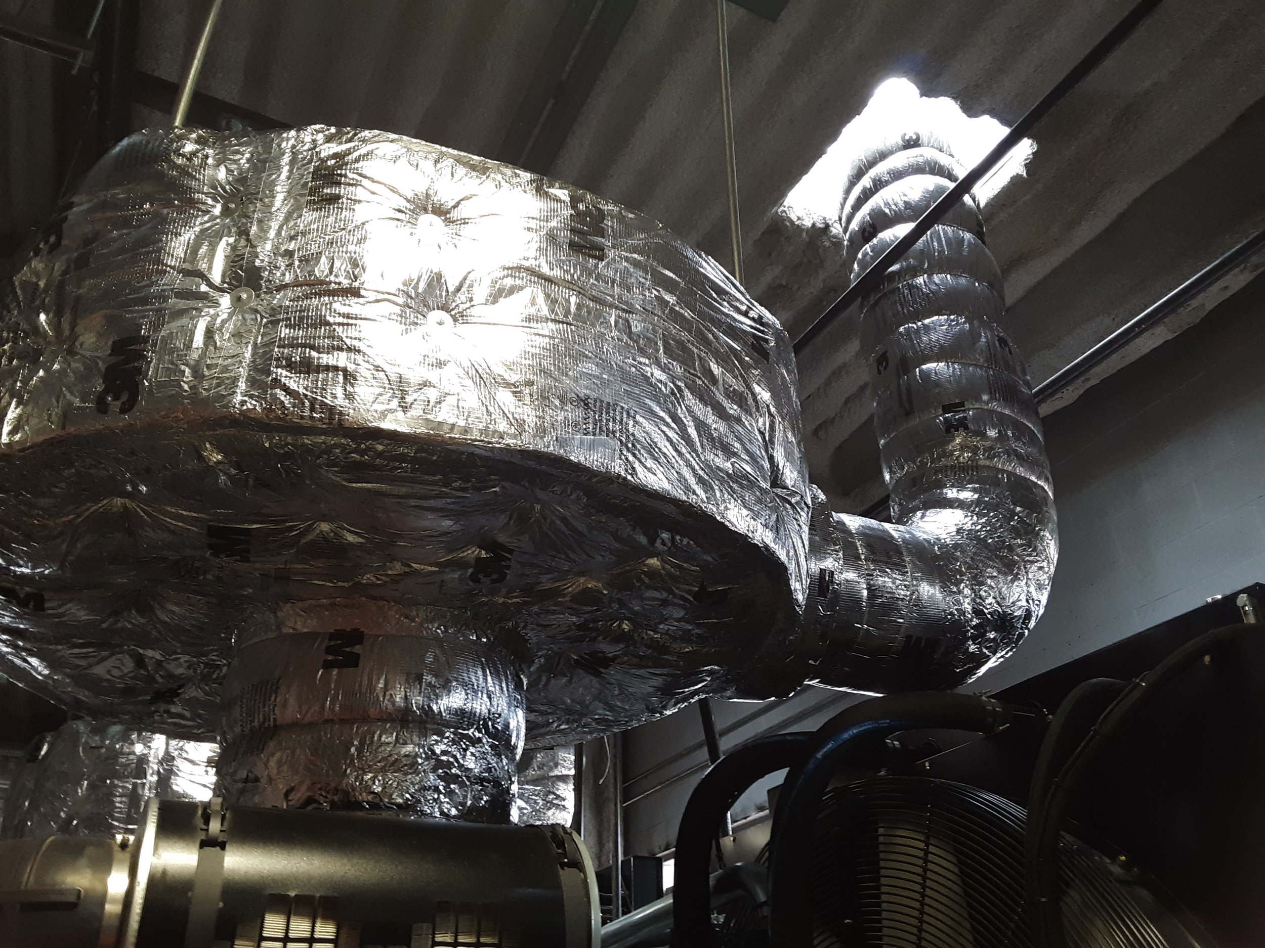 Silver-colored, insulated ductwork running along a ceiling with a hole in the concrete ceiling letting in natural light. Black hoses and a metal fan are visible in the foreground.