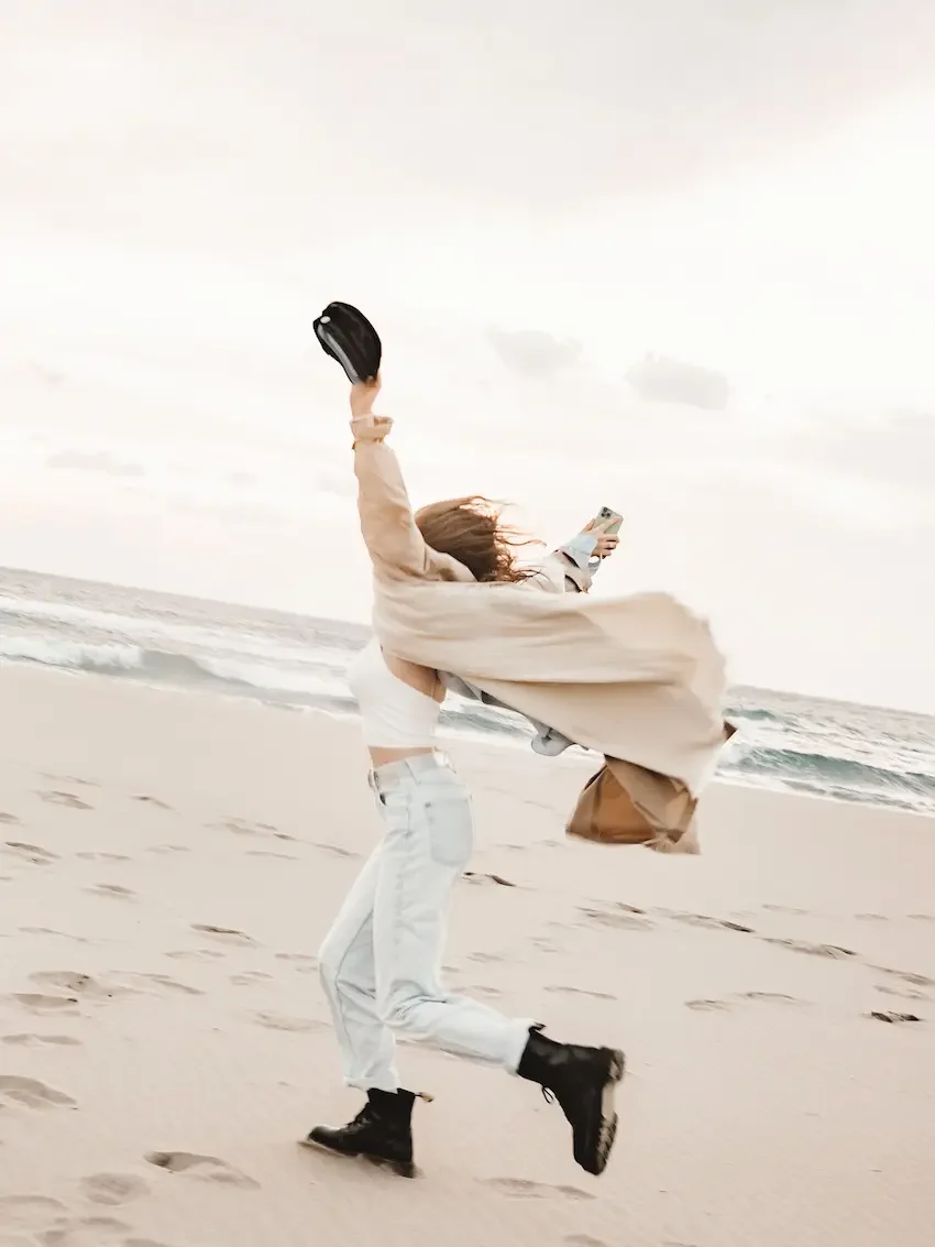 female-in-brown-boots-jeans-and-tan-coat-running-on-beach-with-arms-up-holding-black-cap-in-hand