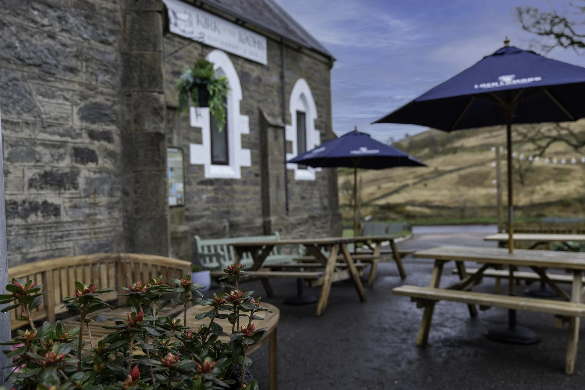 Kirk o'The Lochs outdoor seating area with wooden picnic tables and blue umbrellas, adjacent to a stone building with arched windows. Red-budded plants and a bench are in the foreground, with a grassy hillside in the background.
