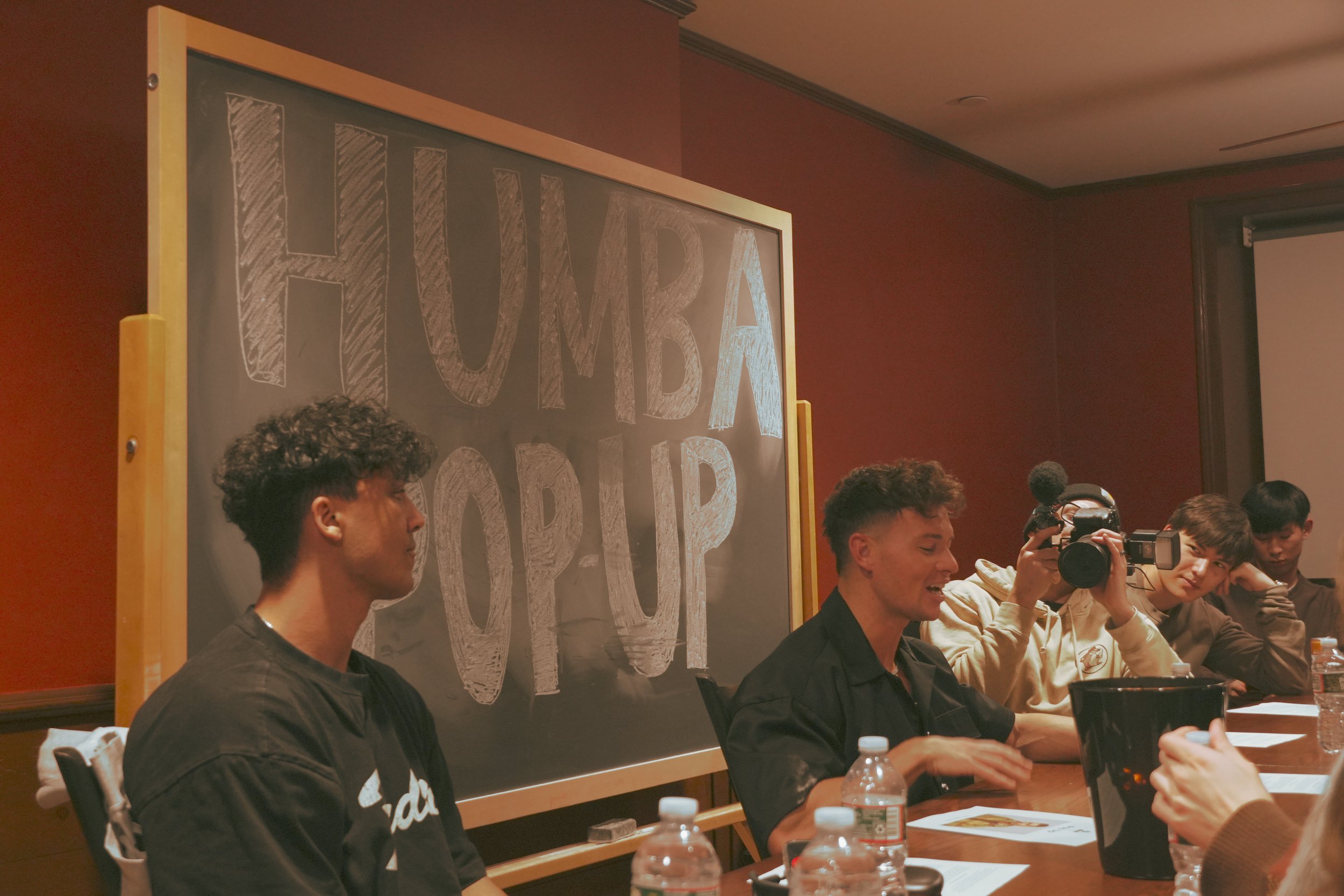 Group of people sitting at a table in front of a chalkboard with 'HUMBA POP-UP' written on it. Several water bottles are on the table.