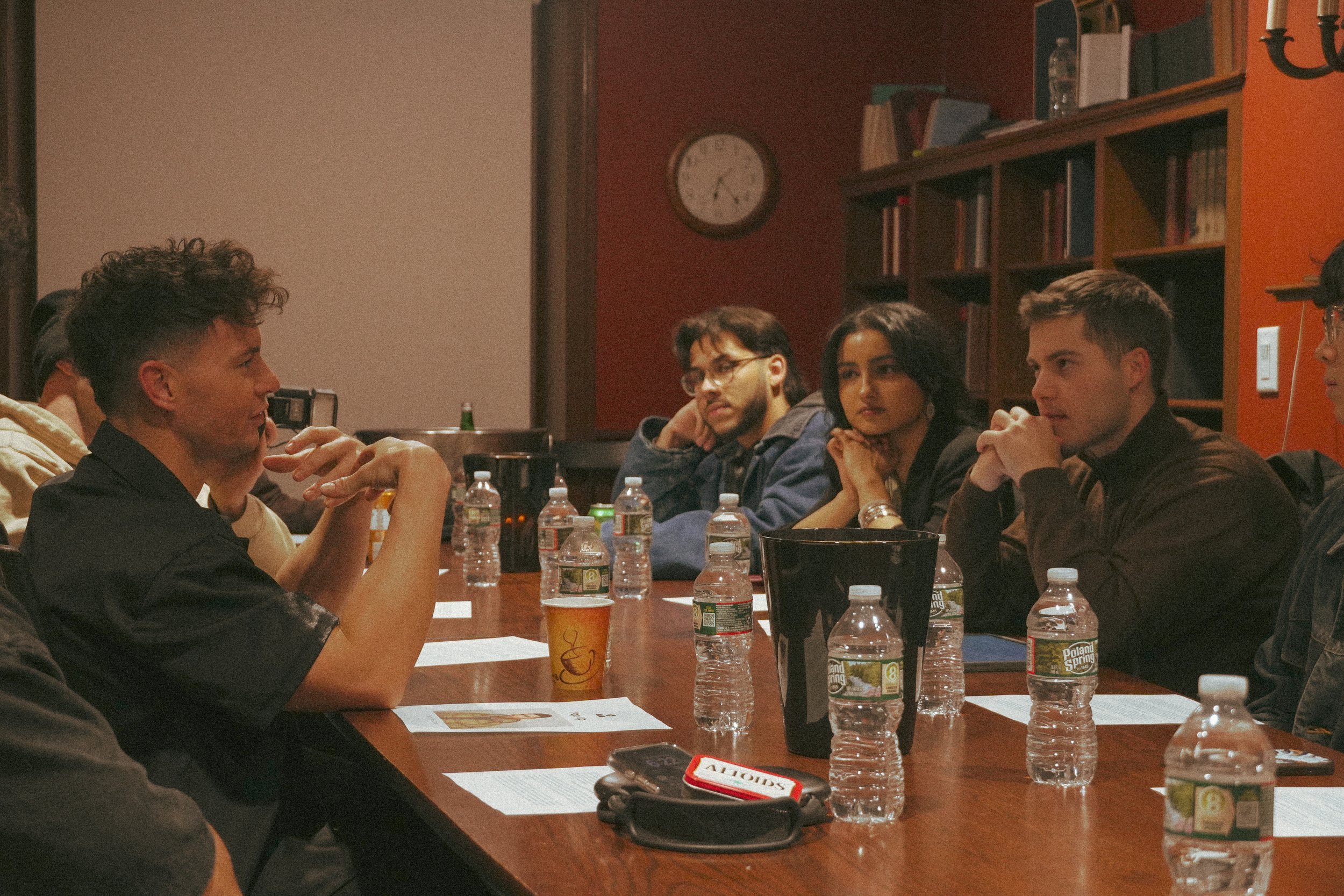 Group of people engaged in a meeting around a conference table, with water bottles and papers in front of them. Bookshelf and clock in background.