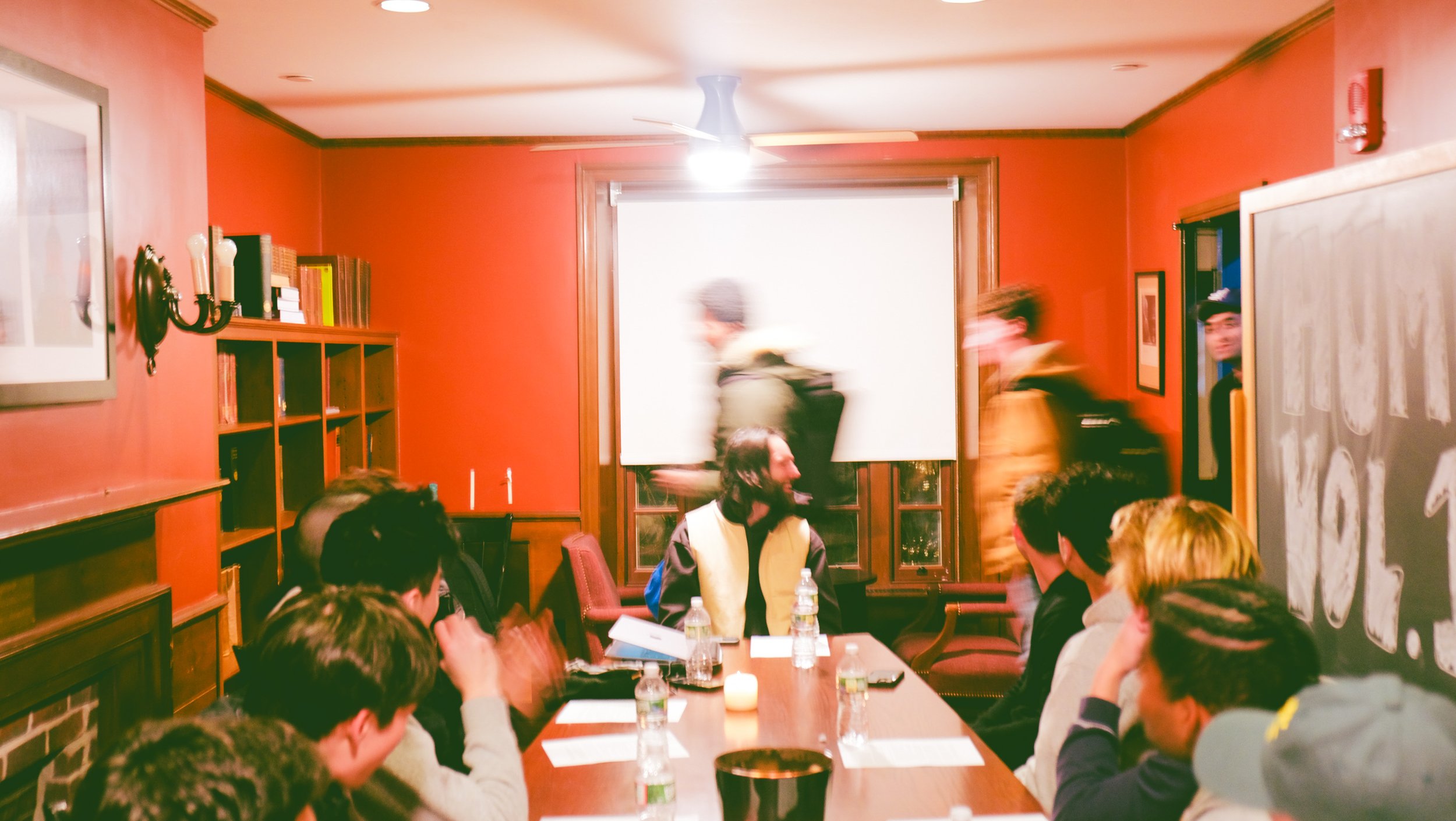 Group of people in a meeting room with a red wall, chalkboard, and bookshelves